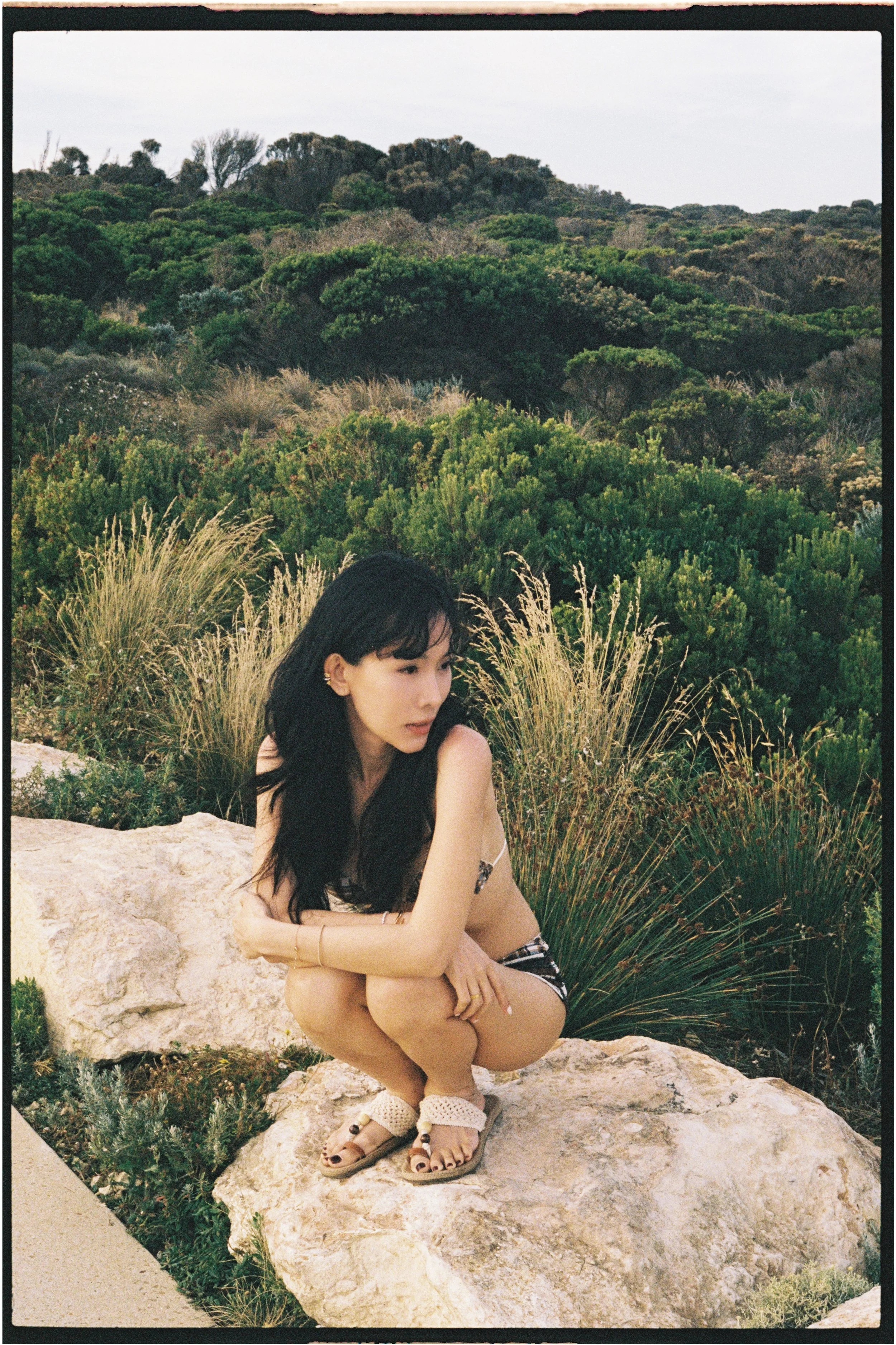 A woman crouching on a large rock in a natural outdoor setting with dense green bushes and trees in the background.