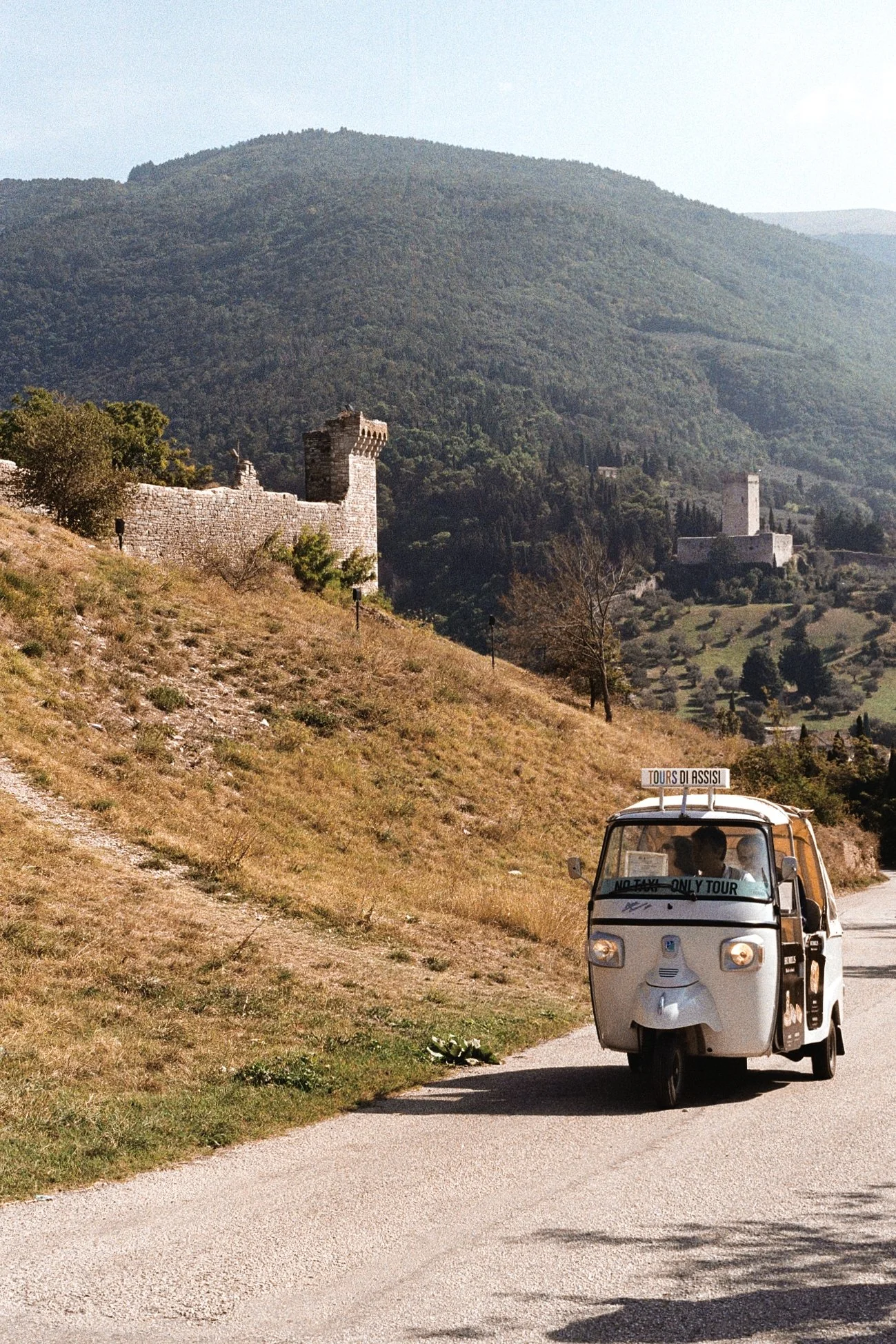 A small, white three-wheeled tour vehicle with a sign on top that reads 'TOURS DI ASSISI' and a banner on the front that says 'NO TAXI - ONLY TOUR,' driving on a narrow road through a hilly landscape with medieval stone castles and green trees in the background.