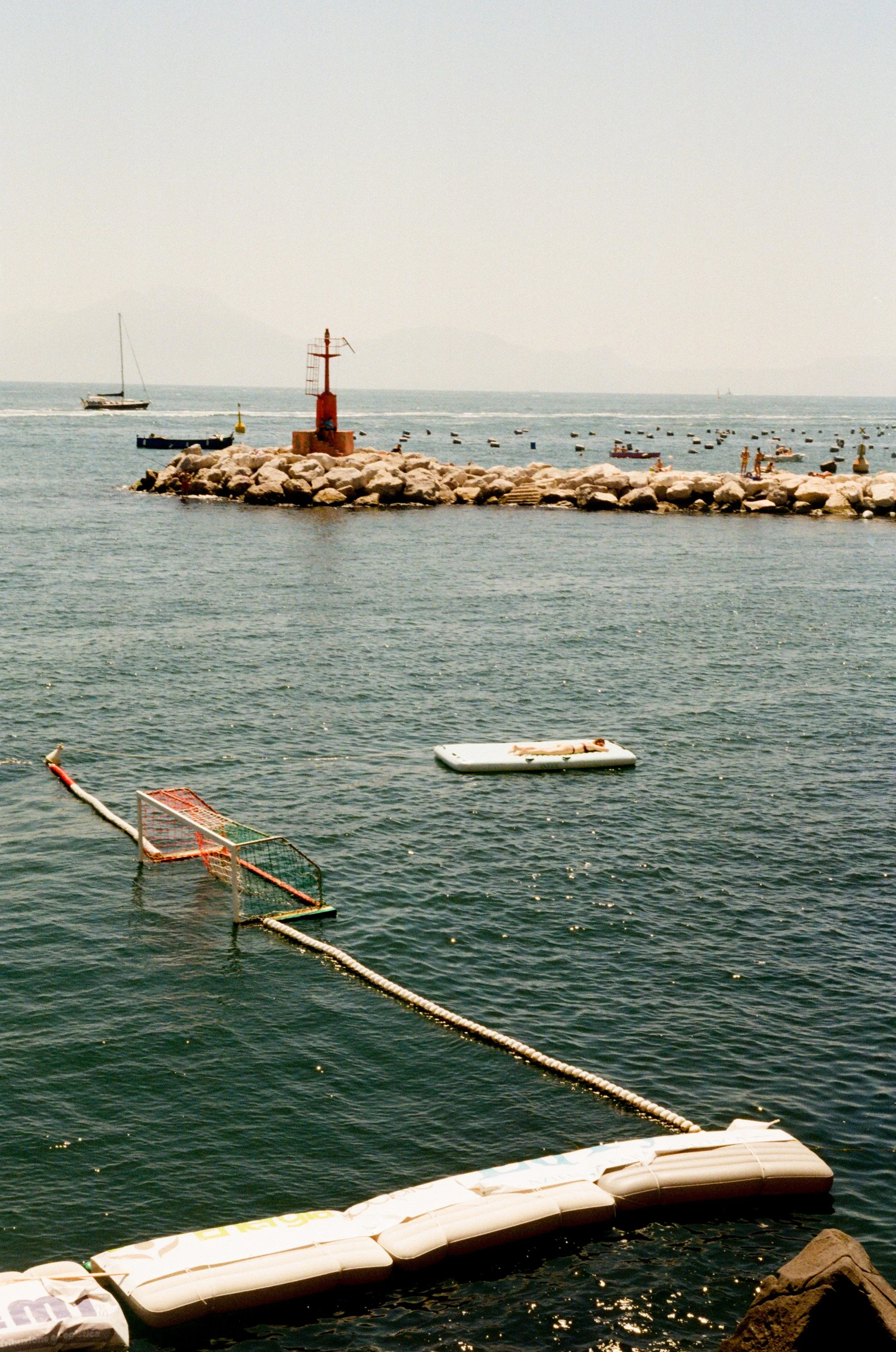 A tranquil harbor scene with a rocky jetty, small boats, a buoyed swimming area, and a distant sailboat, overcast sky, calm waters.