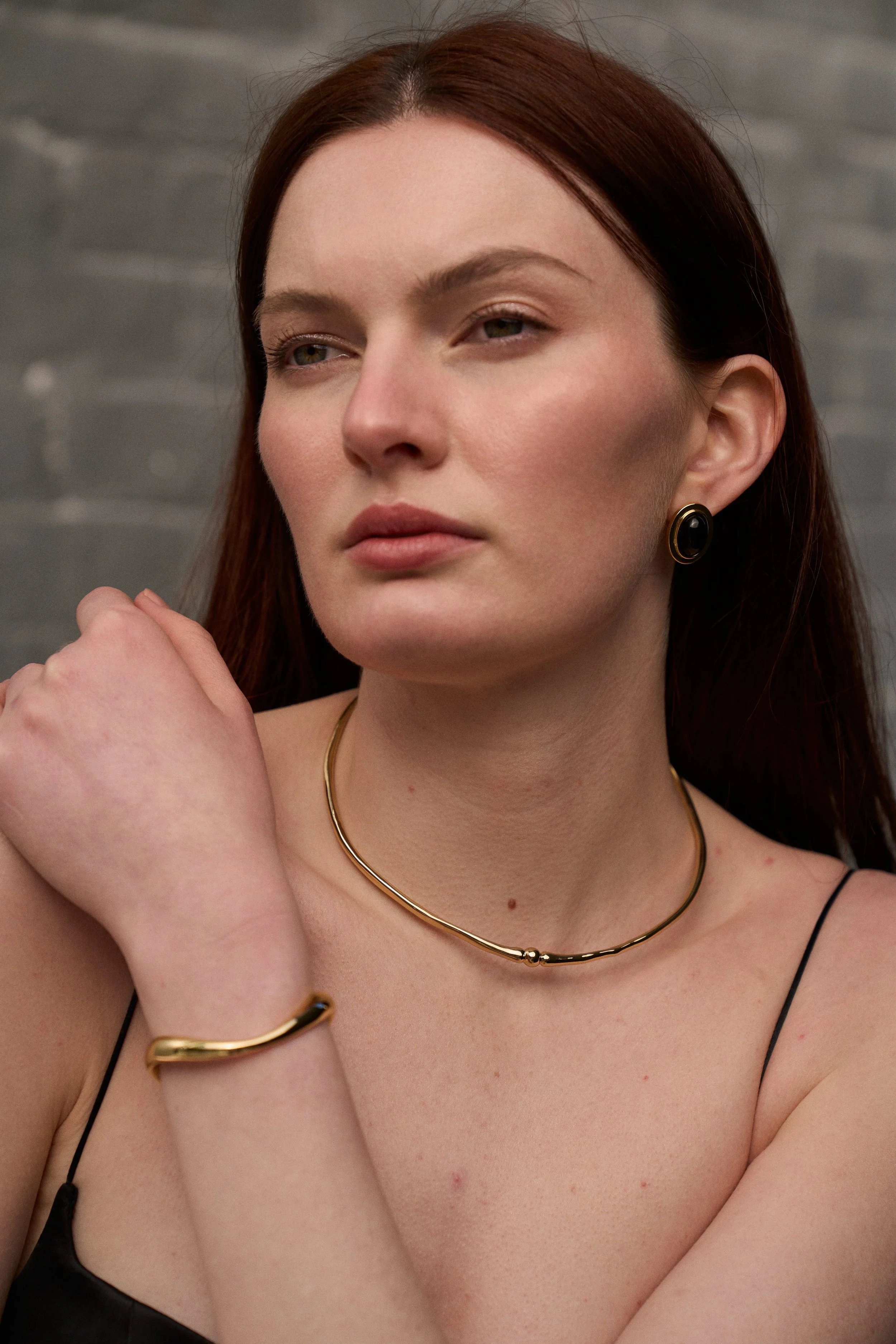 A woman with red hair and fair skin wearing a black spaghetti strap top, gold jewelry including a necklace, bracelet, and earrings, posing against a gray brick wall.