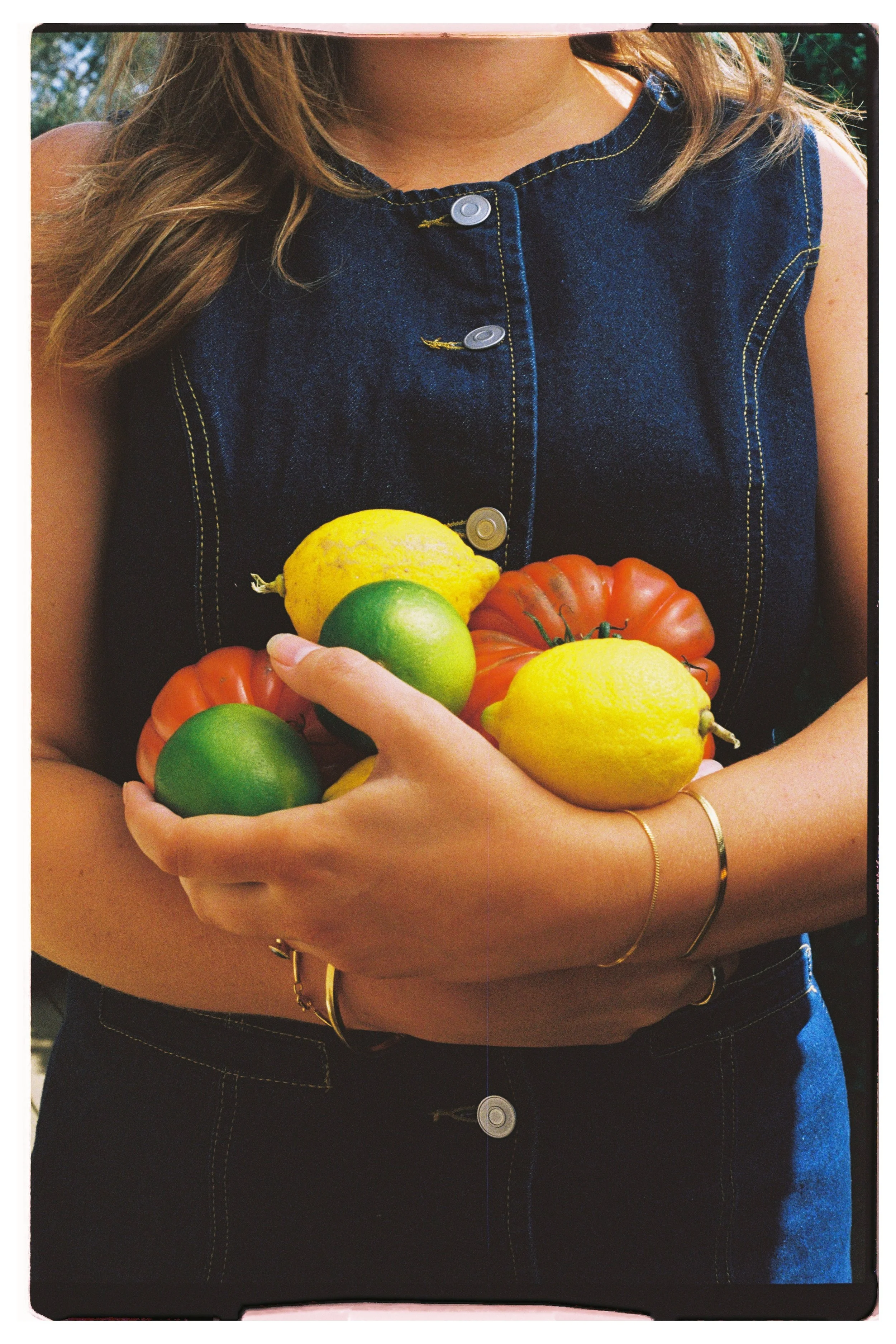 Person holding a handful of fresh vegetables and citrus fruits, including tomatoes, lemons, and limes, wearing a sleeveless denim top with yellow stitching.
