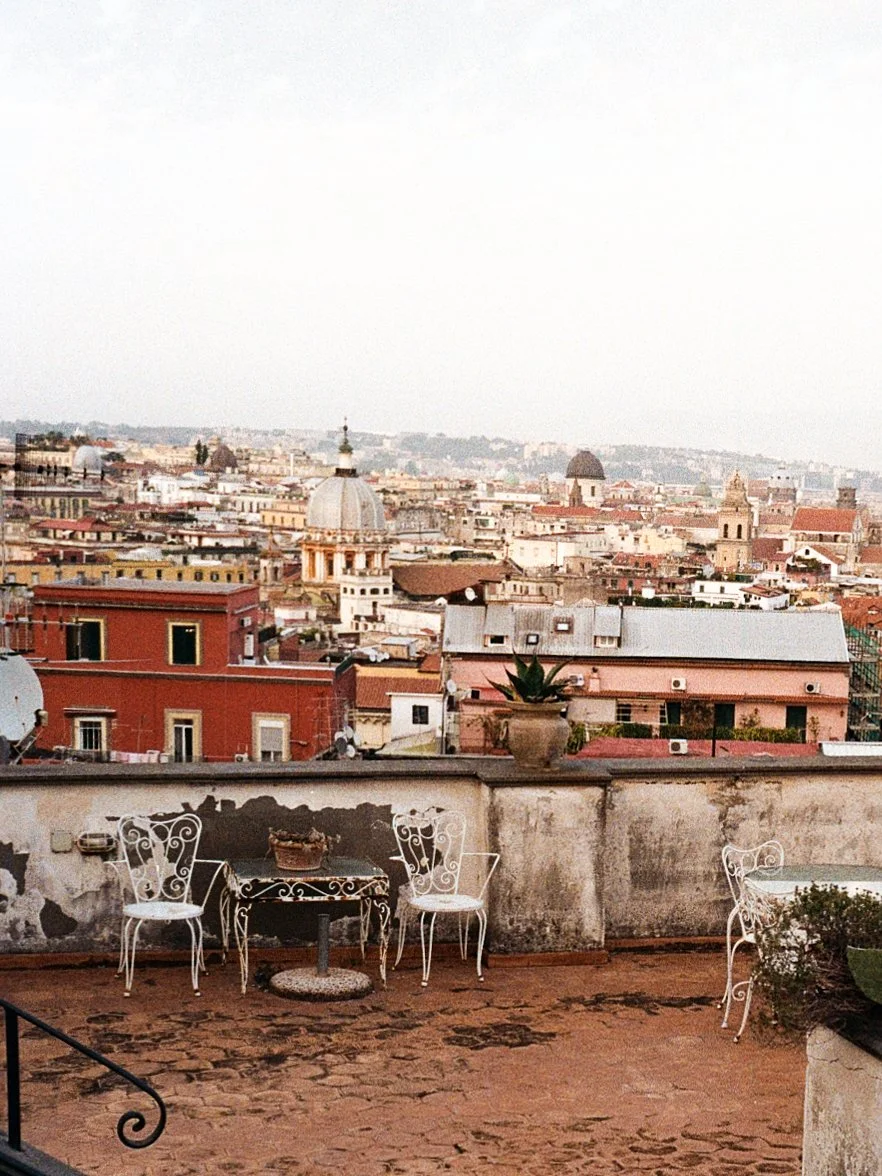 View of a cityscape from a terrace with white iron chairs and a small round table, potted plants, and a weathered wall in the foreground, with historic buildings and domes in the background.