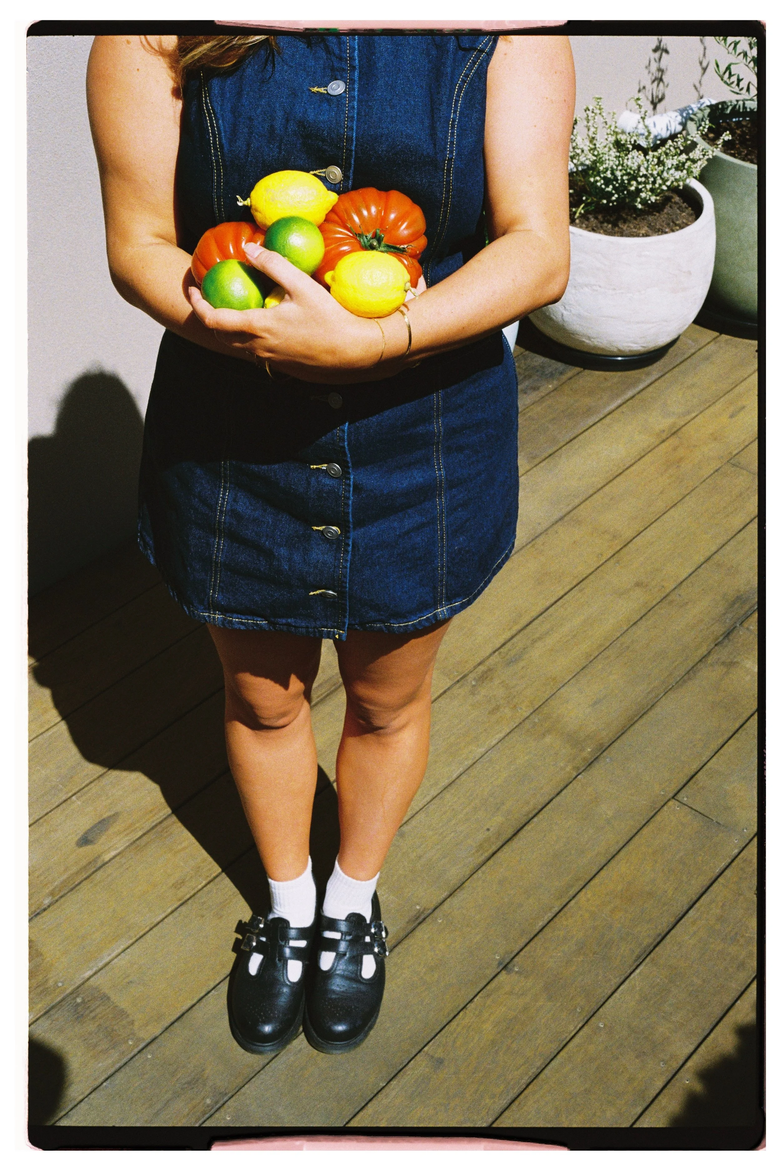 A person wearing a denim dress holds a bowl of tomatoes, lemons, and green apples. They stand on a wooden deck with potted plants in the background.