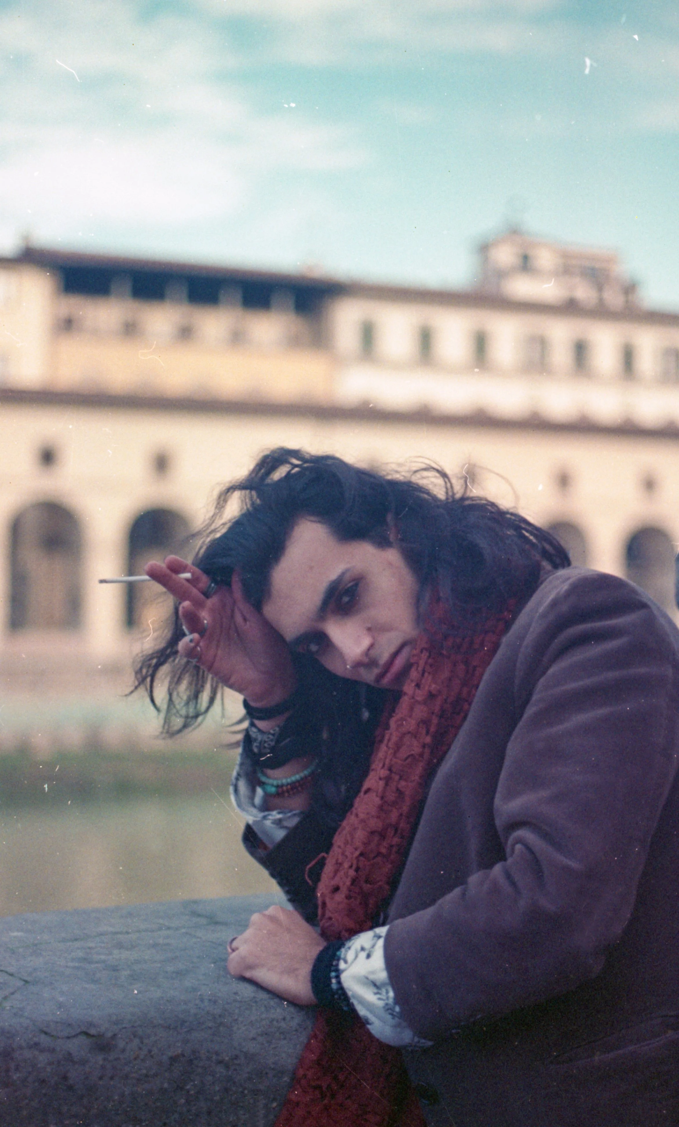 A woman with dark, curly hair leaning on a concrete ledge, holding a cigarette and looking at the camera with a serious expression, with a blurry ancient building with arches in the background under a cloudy sky.
