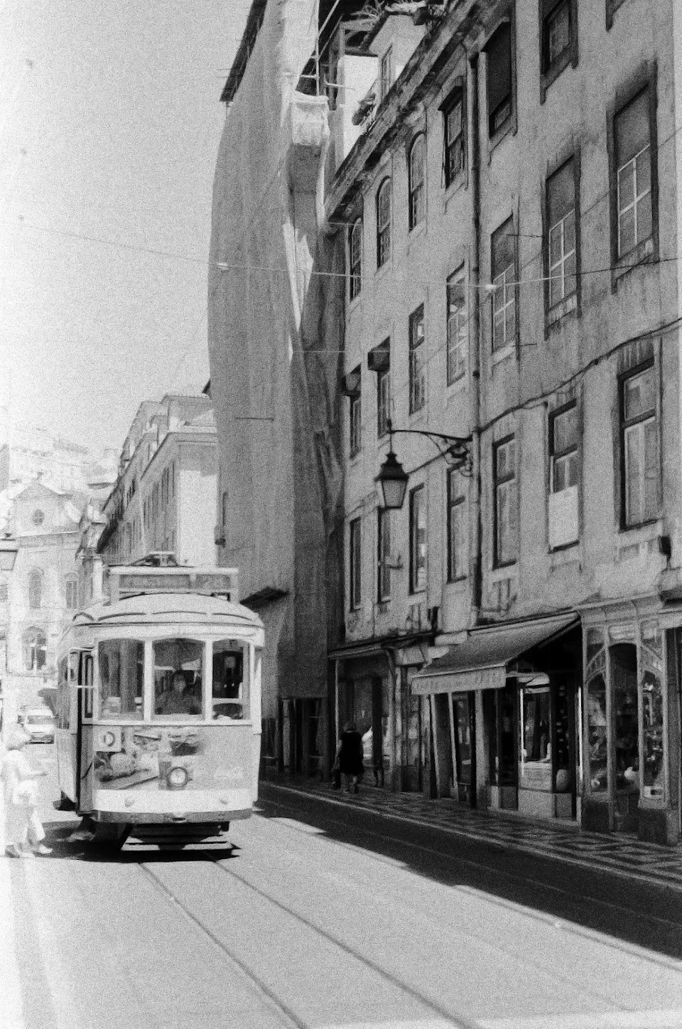 A black and white photo of a city street with a tram car moving along the tracks, pedestrians walking on the sidewalk, and old buildings lining the street. There are street lamps attached to the buildings.