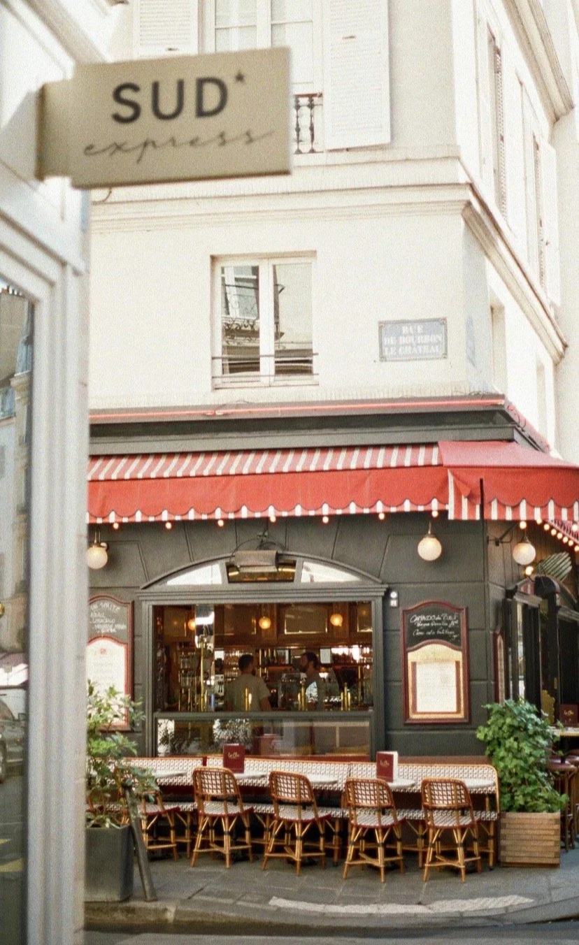 French cafe with red and white striped awning, outdoor seating with chairs, and a view into the inside of the cafe. "SUD express" sign visible in the foreground.