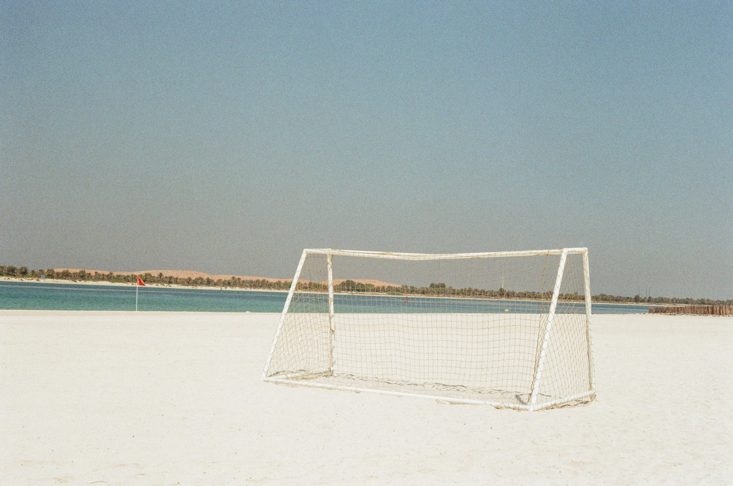 Empty soccer goal on sandy beach with water and distant land in the background.
