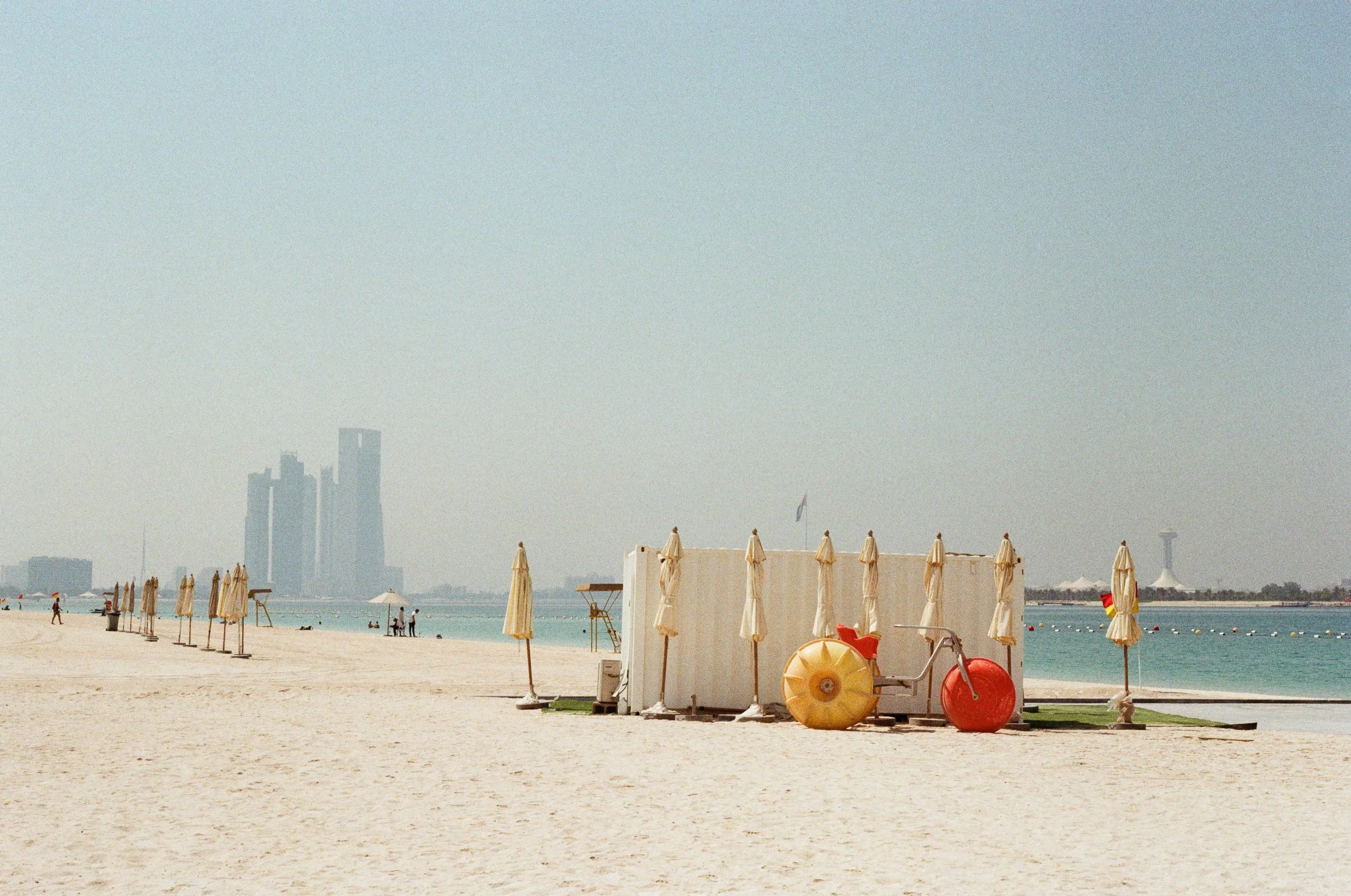 Empty beach with closed umbrellas, a lifeguard station, and a city skyline in the distance.