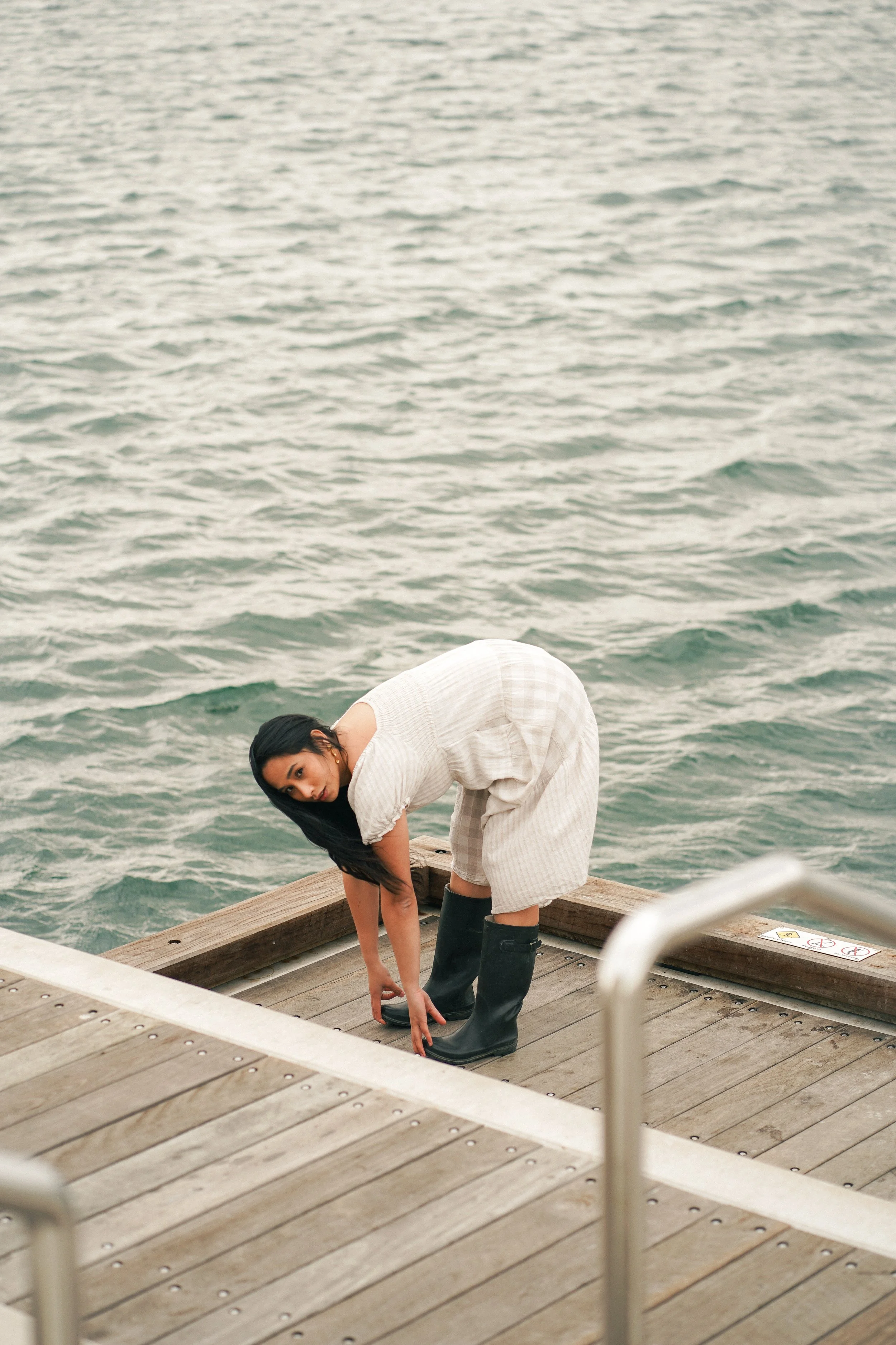Woman in light-colored dress and black boots bending down on a wooden dock near water, adjusting her shoe.