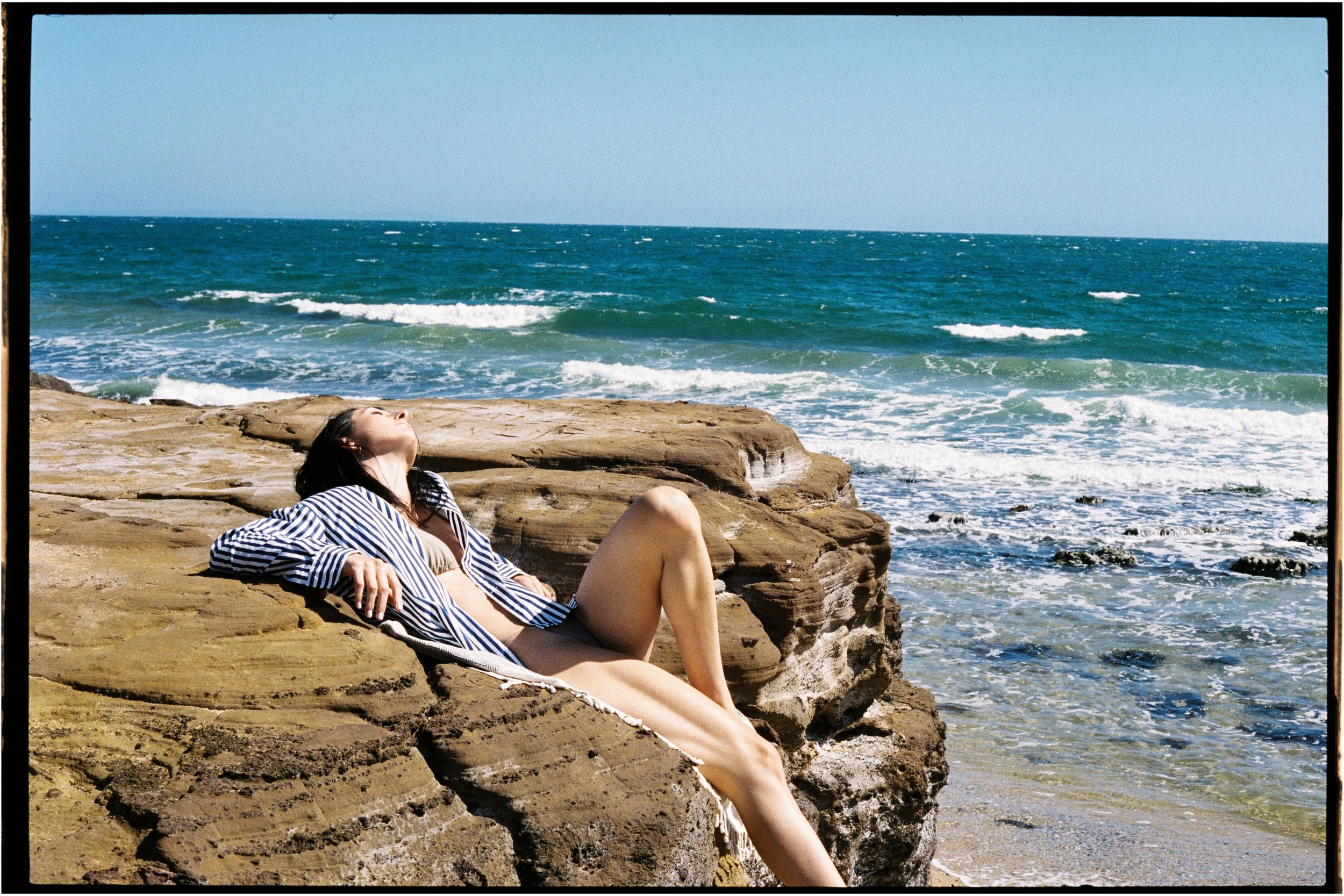 A woman relaxing on a rocky beach, sunbathing near the ocean with waves and clear blue sky in the background.
