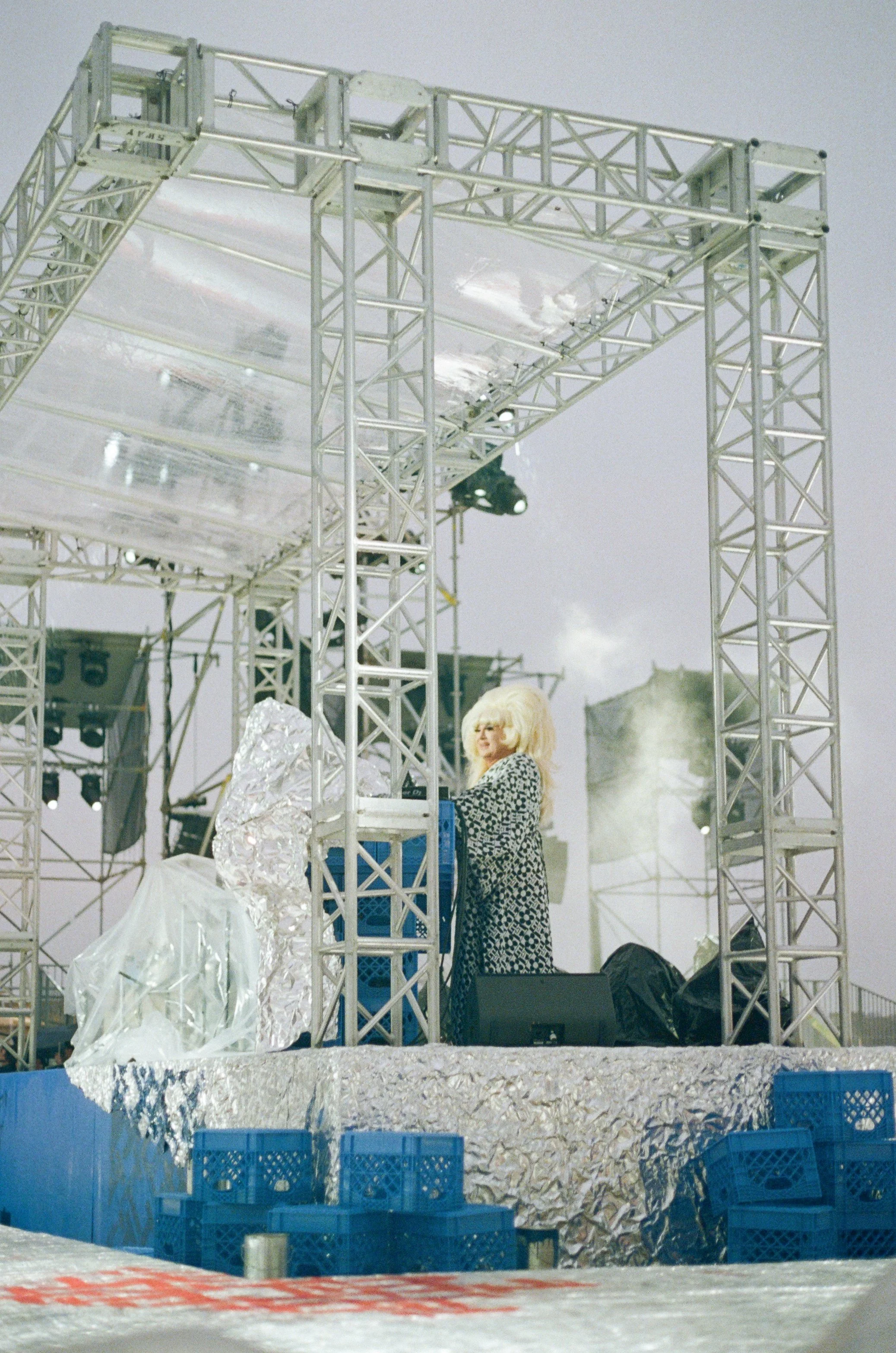 A woman with blonde hair wearing a patterned coat stands at a DJ booth on an outdoor stage, surrounded by metal scaffolding, plastic crates, and stage equipment, with a cloudy sky in the background.