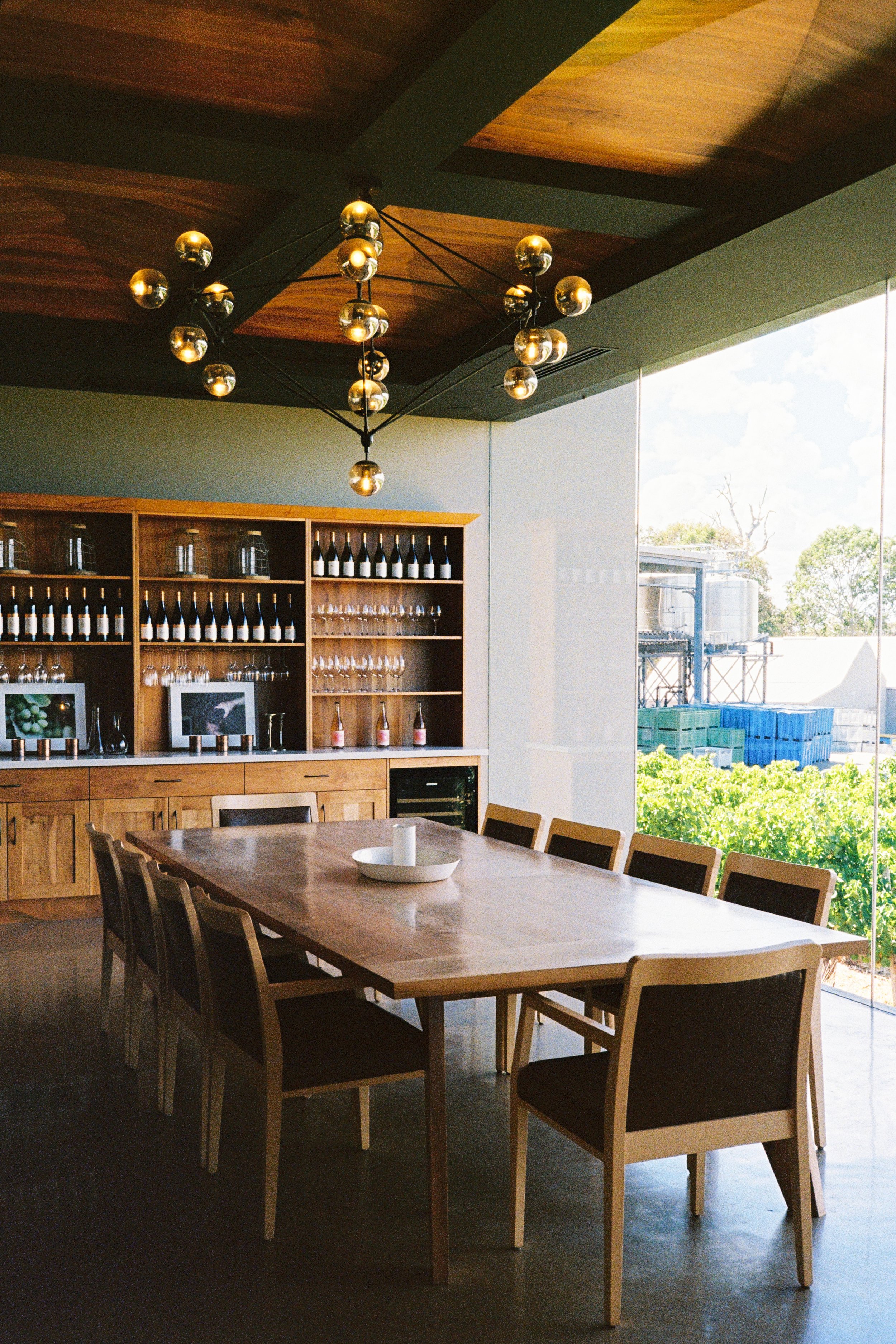 Modern dining room with a large wooden table, nine chairs, a decorative bowl with candles, and a bright view outside through large glass windows. There is a wooden cabinet with wine bottles and glasses, and a stylish chandelier hanging from a wooden ceiling.
