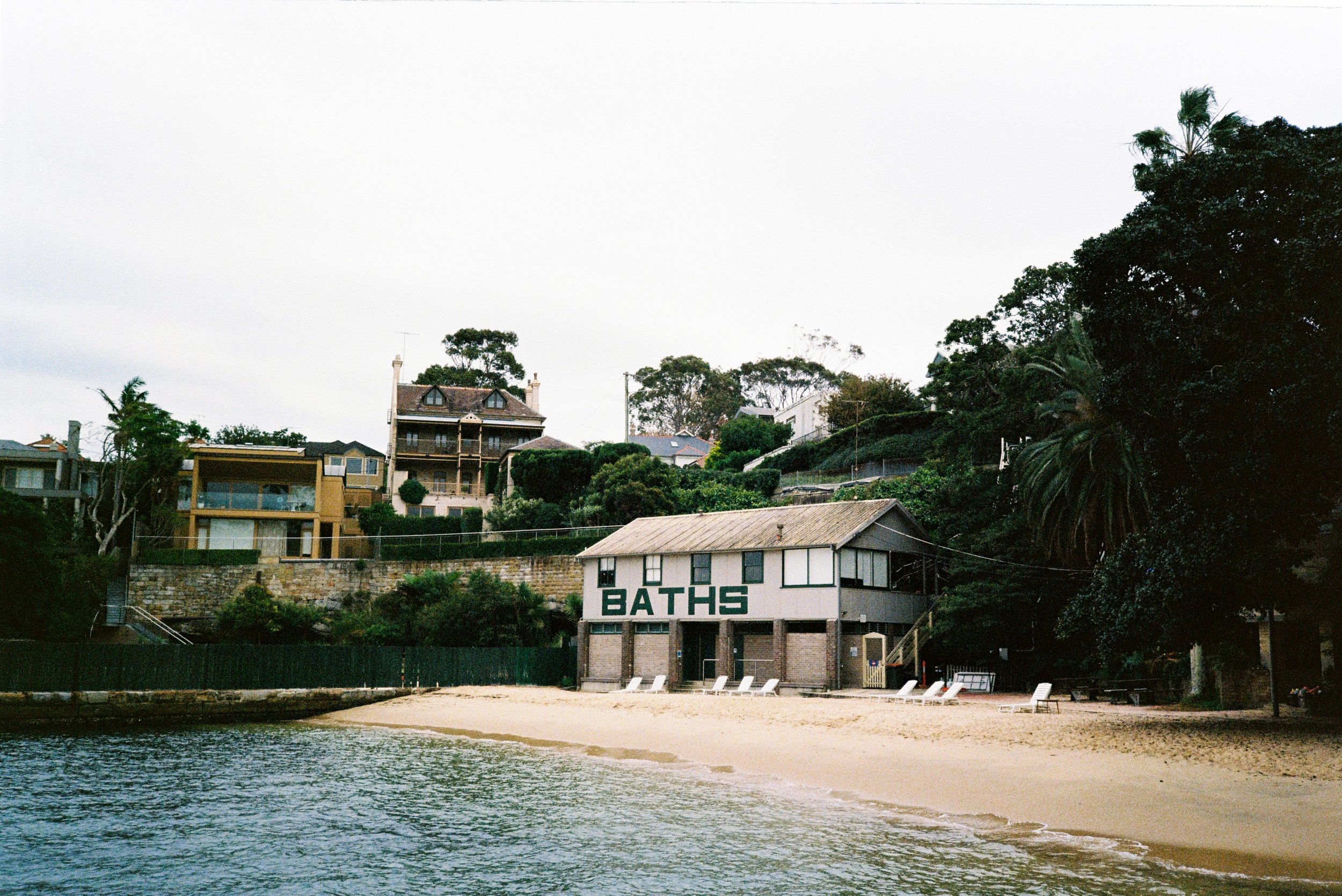 Beach scene with a sandy shore, water, trees, and houses in the background; a building labeled 'BATHS' on the beach.