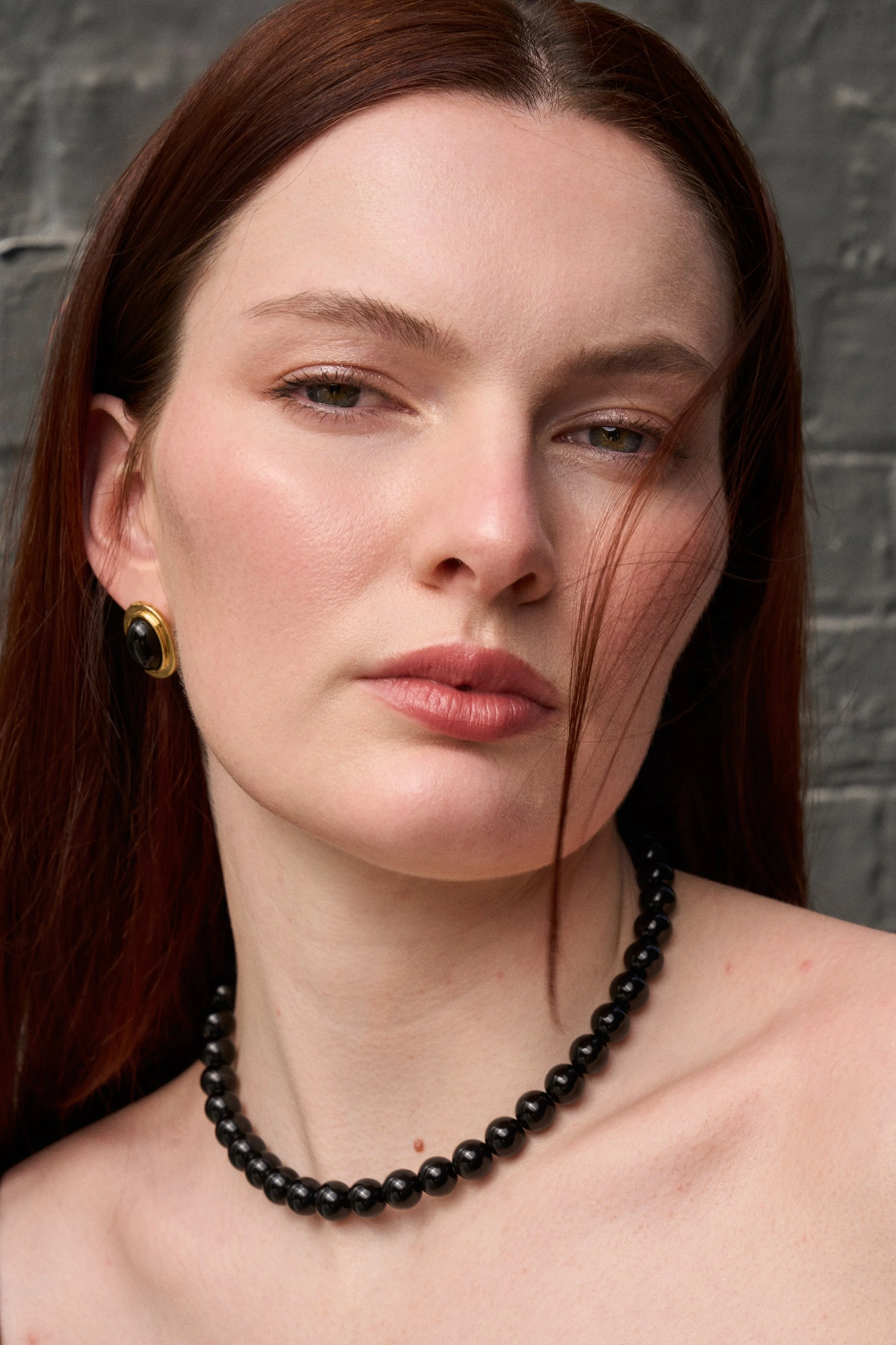 A close-up of a woman with red hair, wearing gold and black earrings, a black beaded necklace, and a subtle makeup look, against a gray brick wall background.
