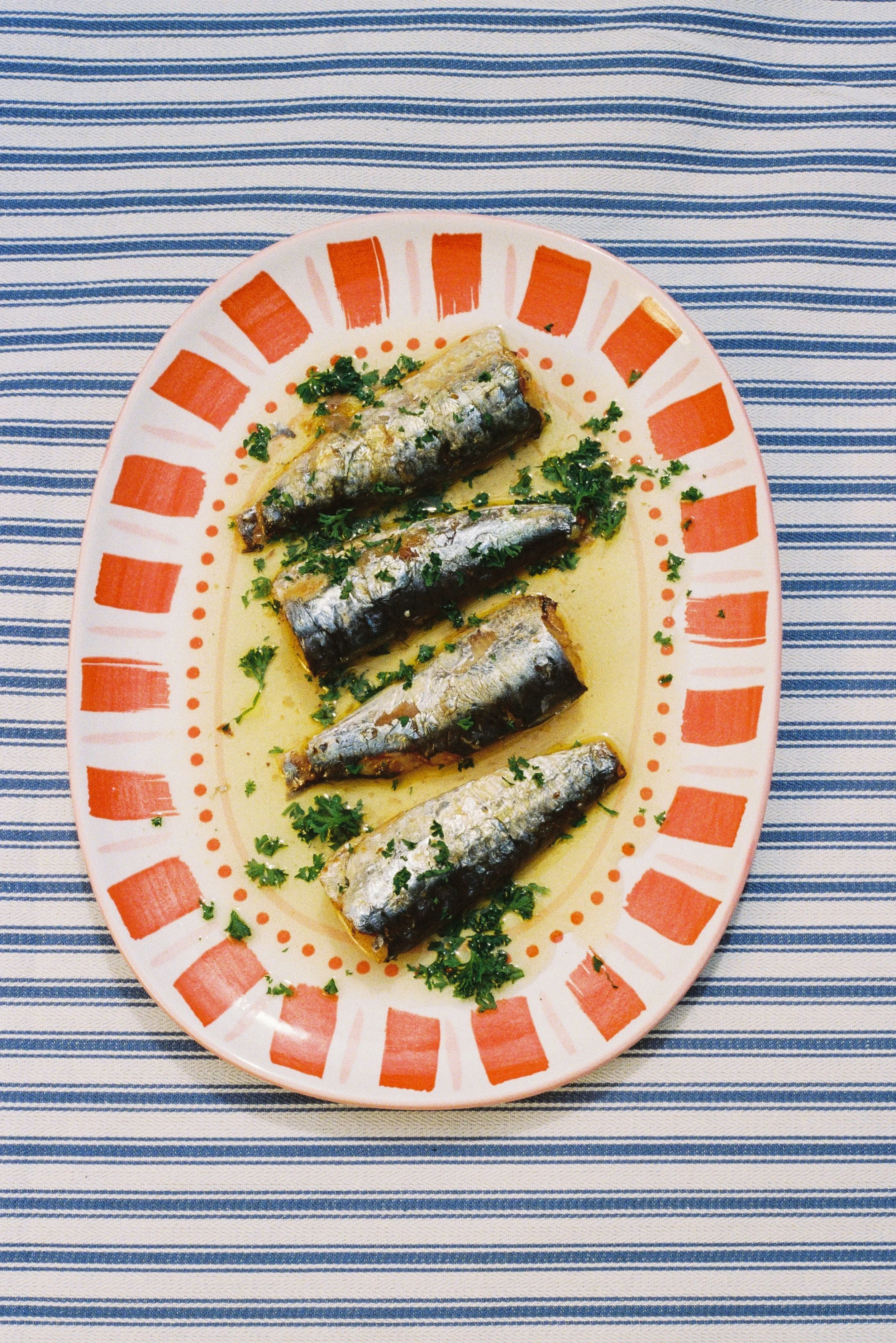 A plate of four cooked sardines garnished with chopped parsley, served in a light sauce, on a decorative orange and white platter, placed on a blue and white striped tablecloth.