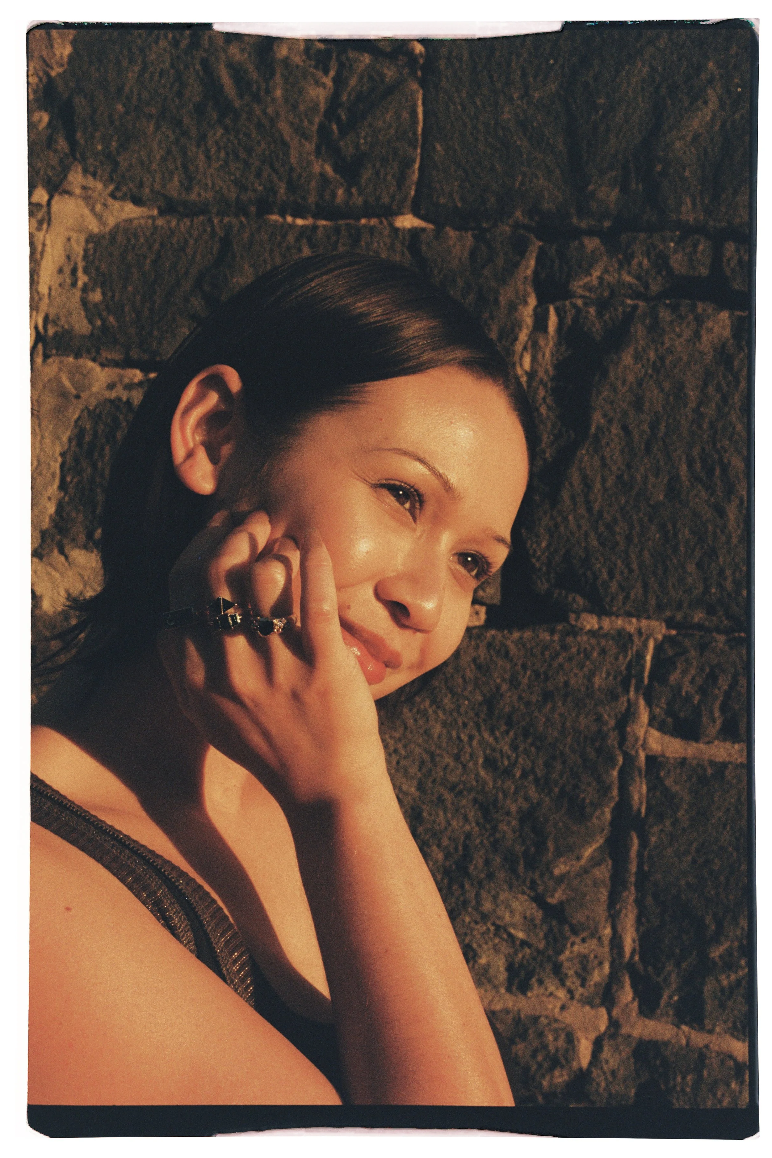 A woman with slicked-back dark hair smiling and posing against a stone wall, illuminated by warm light.