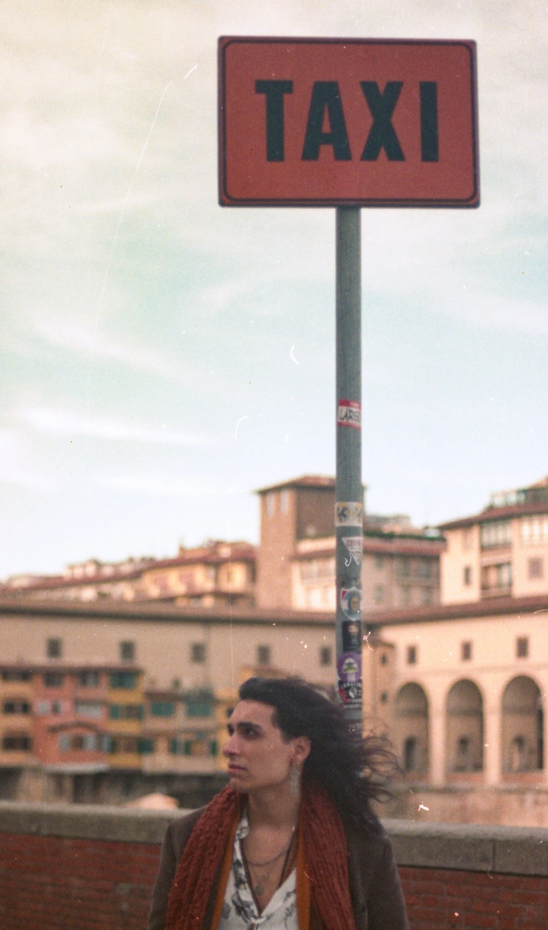 A woman with dark hair and earrings stands outdoors in front of a cityscape, with a red taxi sign on a pole above her.