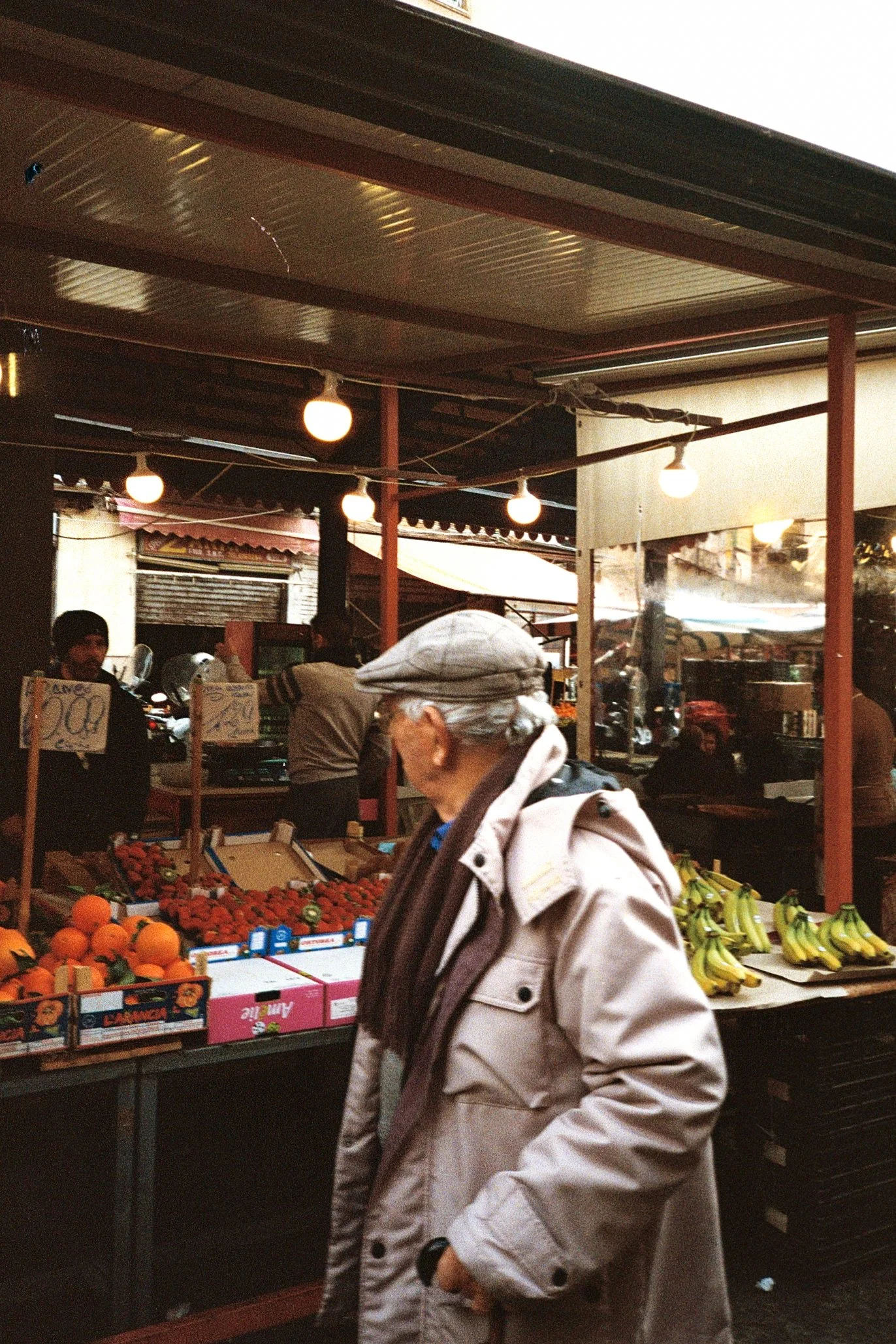 An elderly woman wearing a gray cap and beige jacket is walking past a fruit stand at an outdoor market, with strawberries, bananas, and oranges visible on the stall.