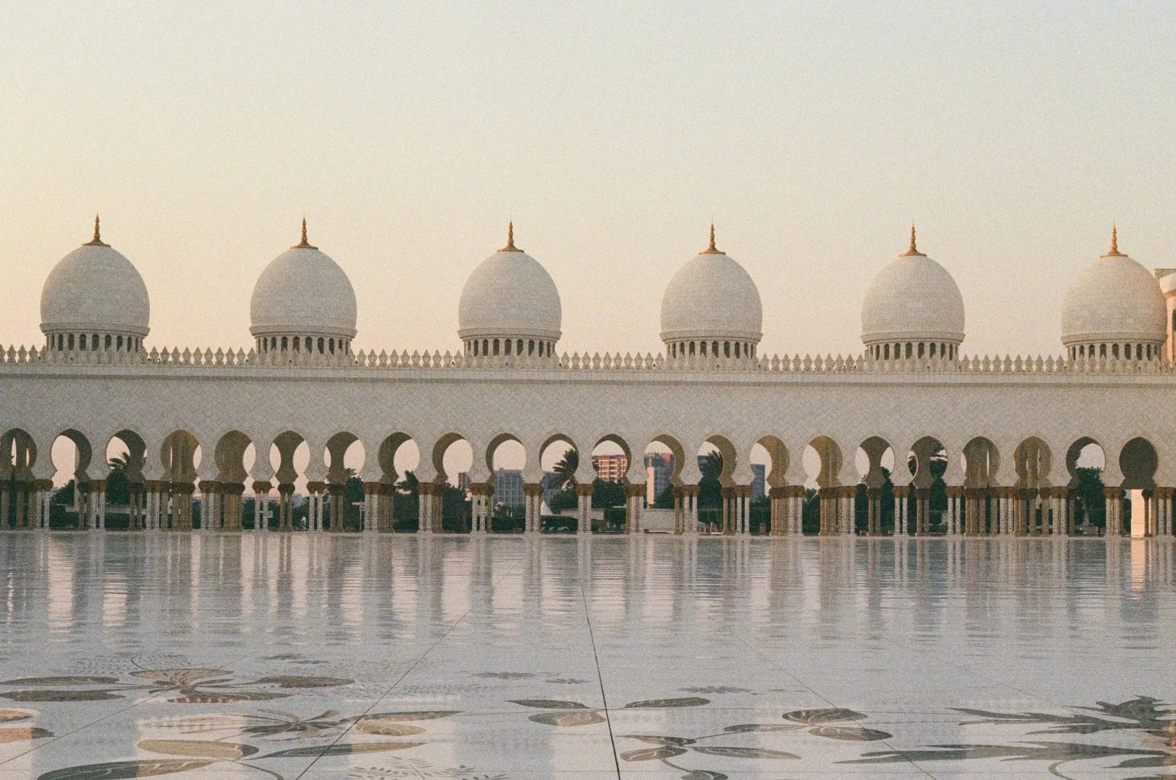 View of a mosque with five white domes, arched openings, and reflection on a polished marble surface.