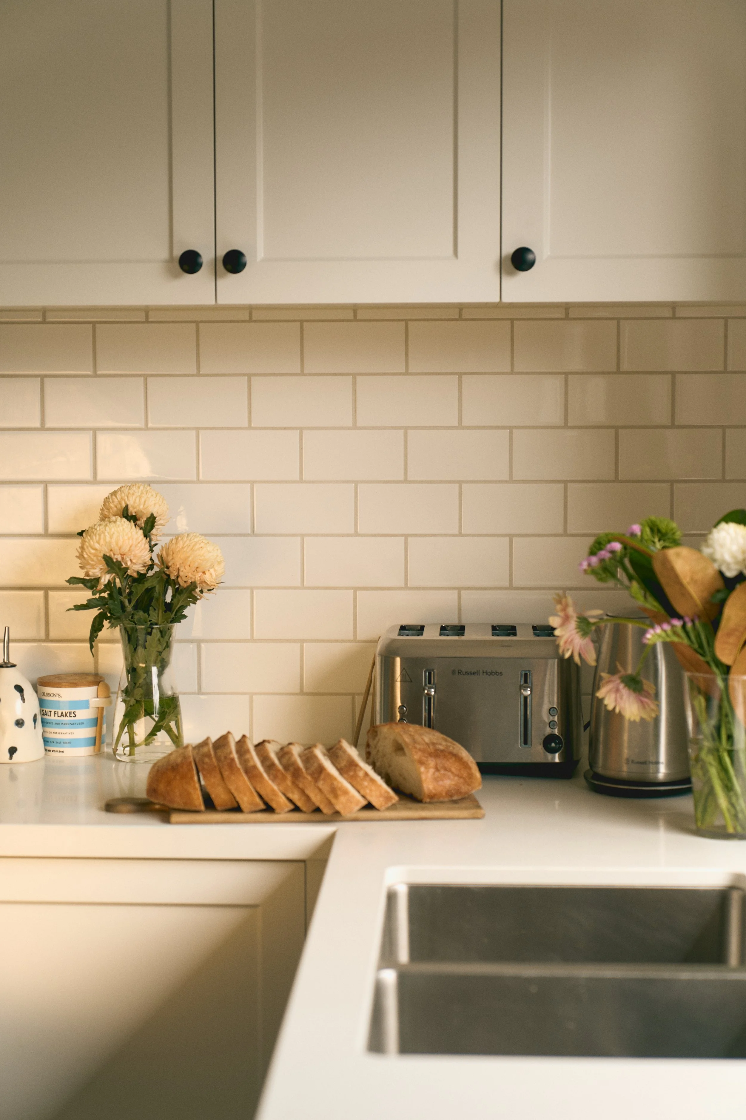 Kitchen countertop with sliced bread on a cutting board, cream-colored cabinets, white subway tile backsplash, a vase with flowers, a stainless steel toaster, and a flower vase.