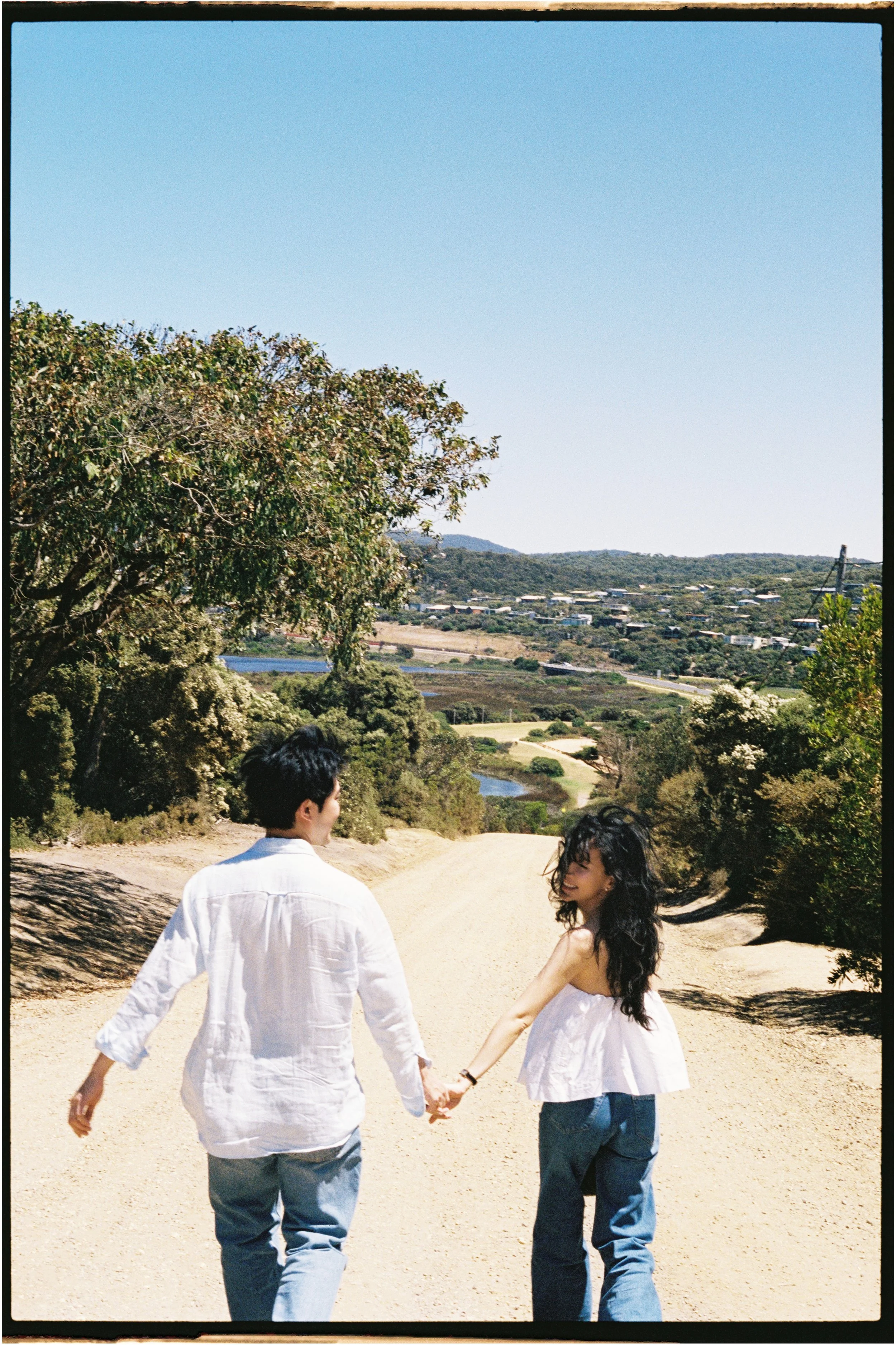 A couple holding hands and walking on a dirt path in a scenic outdoor setting with trees and a distant view of houses and hills under a clear blue sky.
