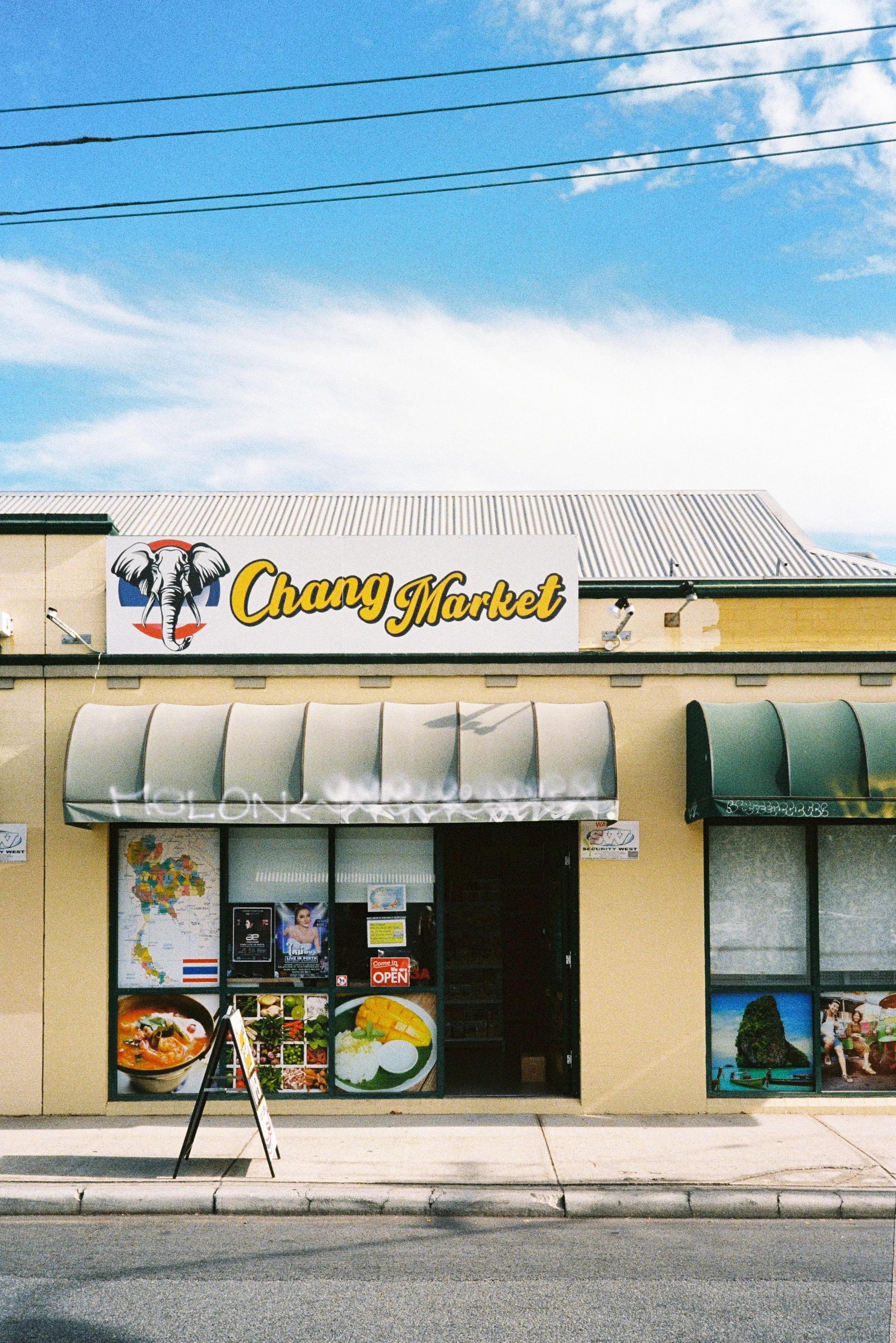 The storefront of Chang Market with an elephant logo, awnings over the windows, and posters displaying food items. There are power lines and a blue sky with clouds above.