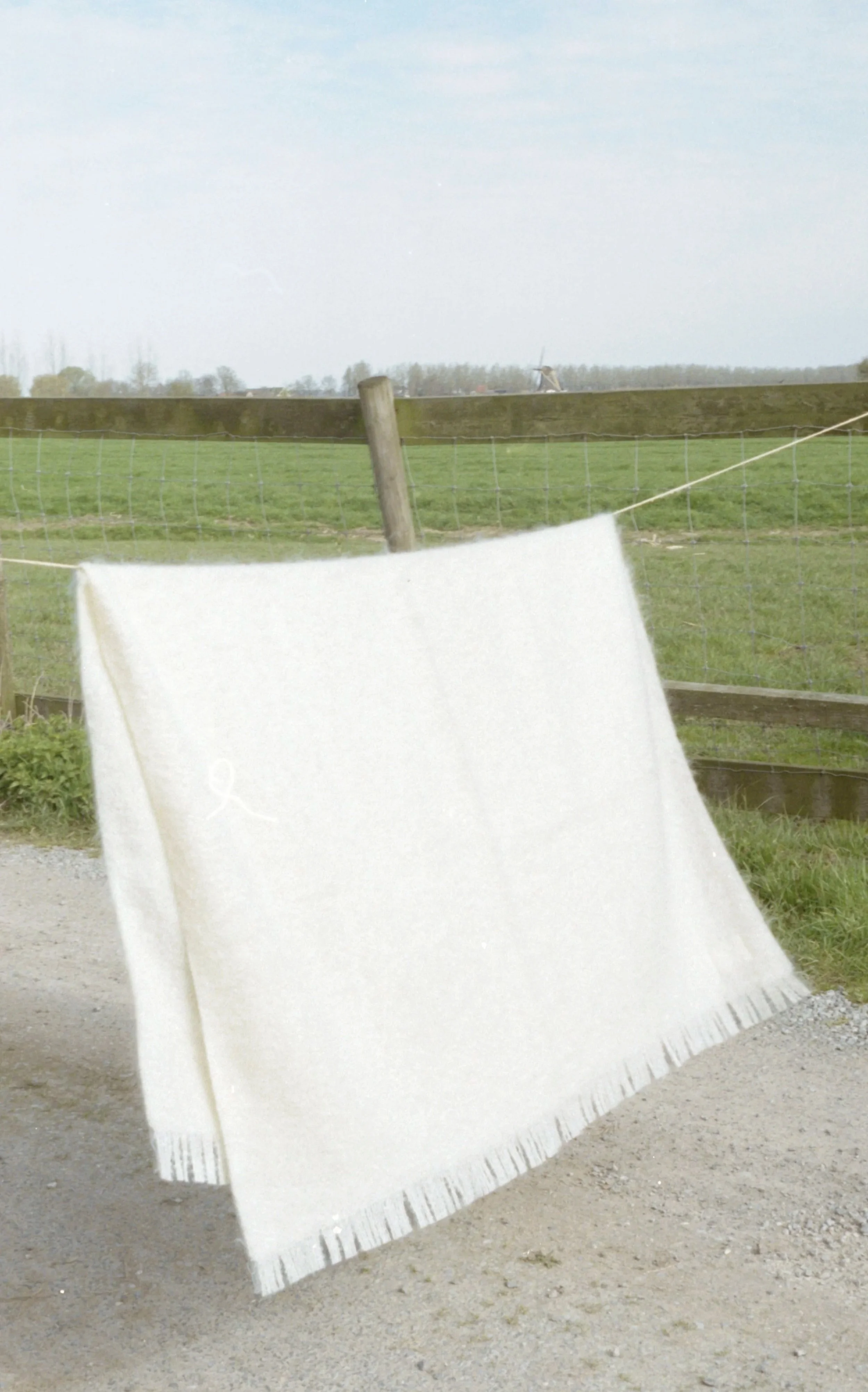 White blanket hanging on a clothesline in a rural outdoor setting with grass, a fence, and distant trees and sky.