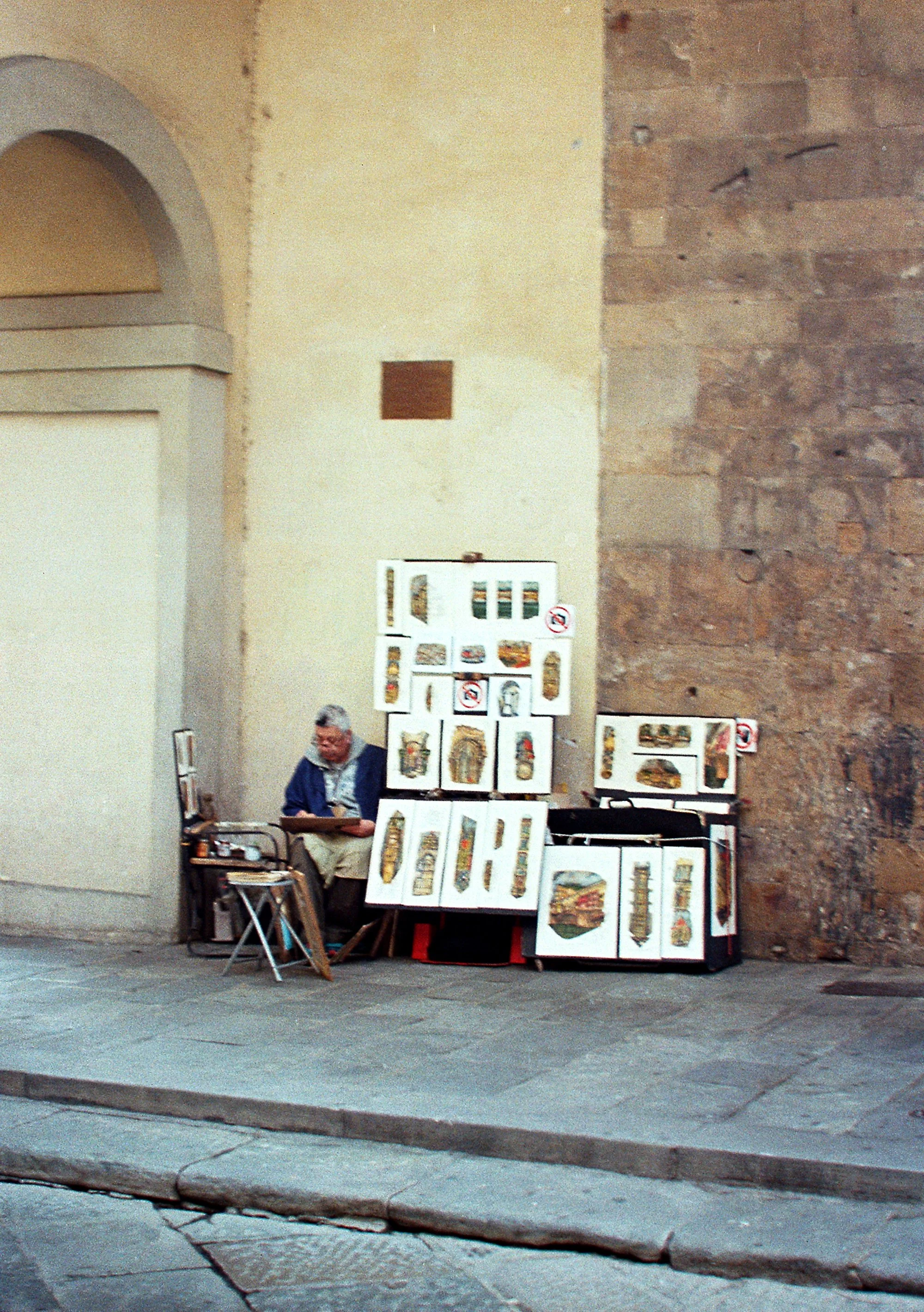An older man sitting on a chair beside a street corner with artistic prints displayed on boards, with a stone and plaster wall behind him.