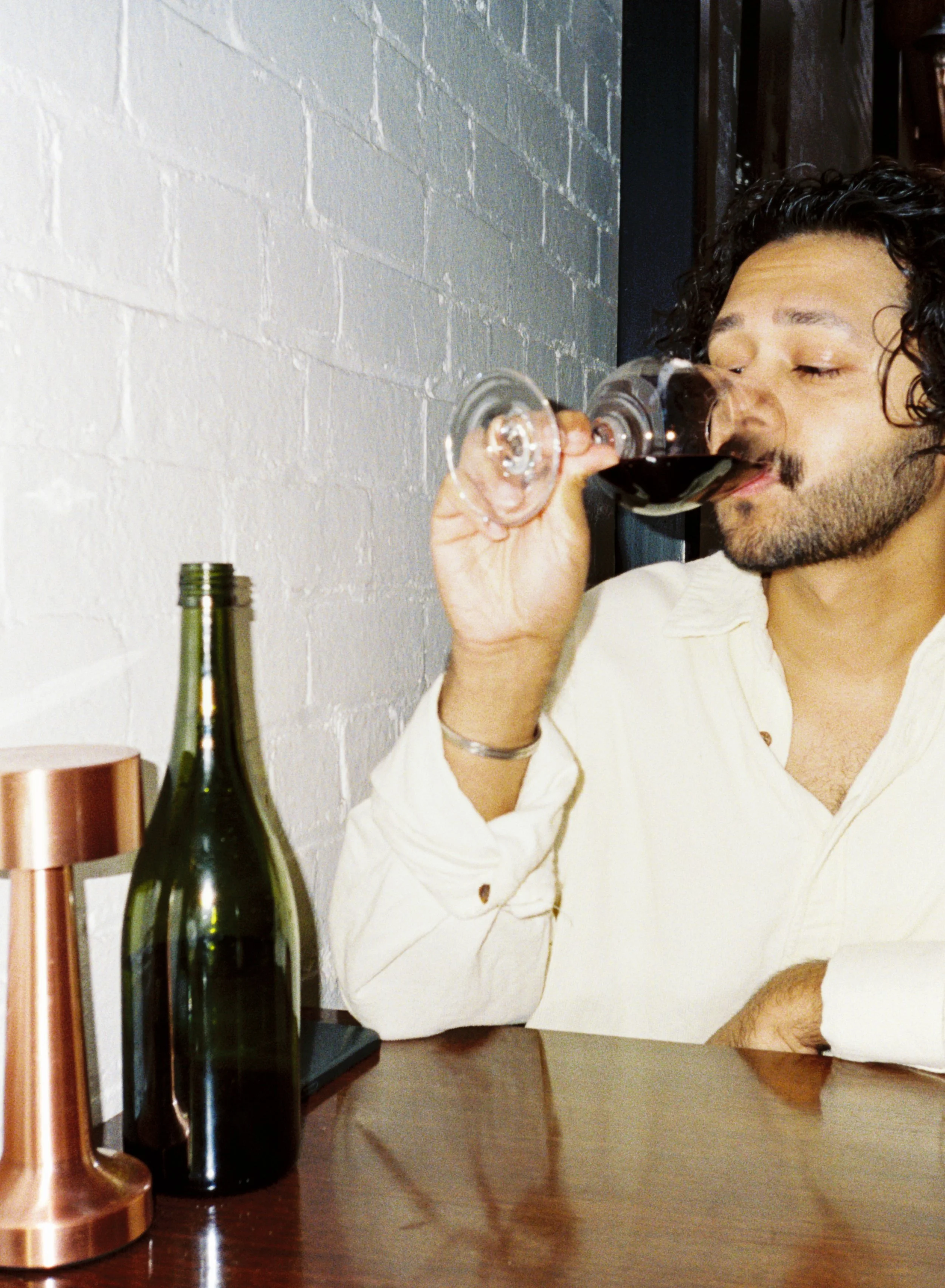 A man with curly hair and a beard is sitting at a table, drinking red wine from a wine glass.