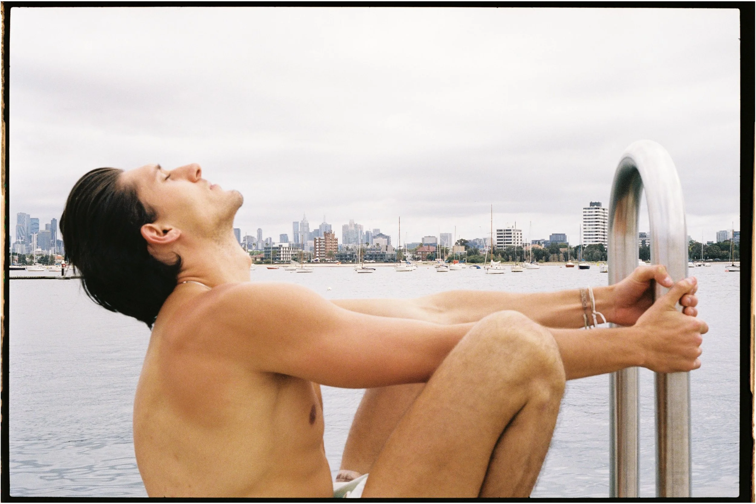 A shirtless man sitting on a dock with his knees drawn up, holding a metal ladder with a city skyline and boats on the water in the background.