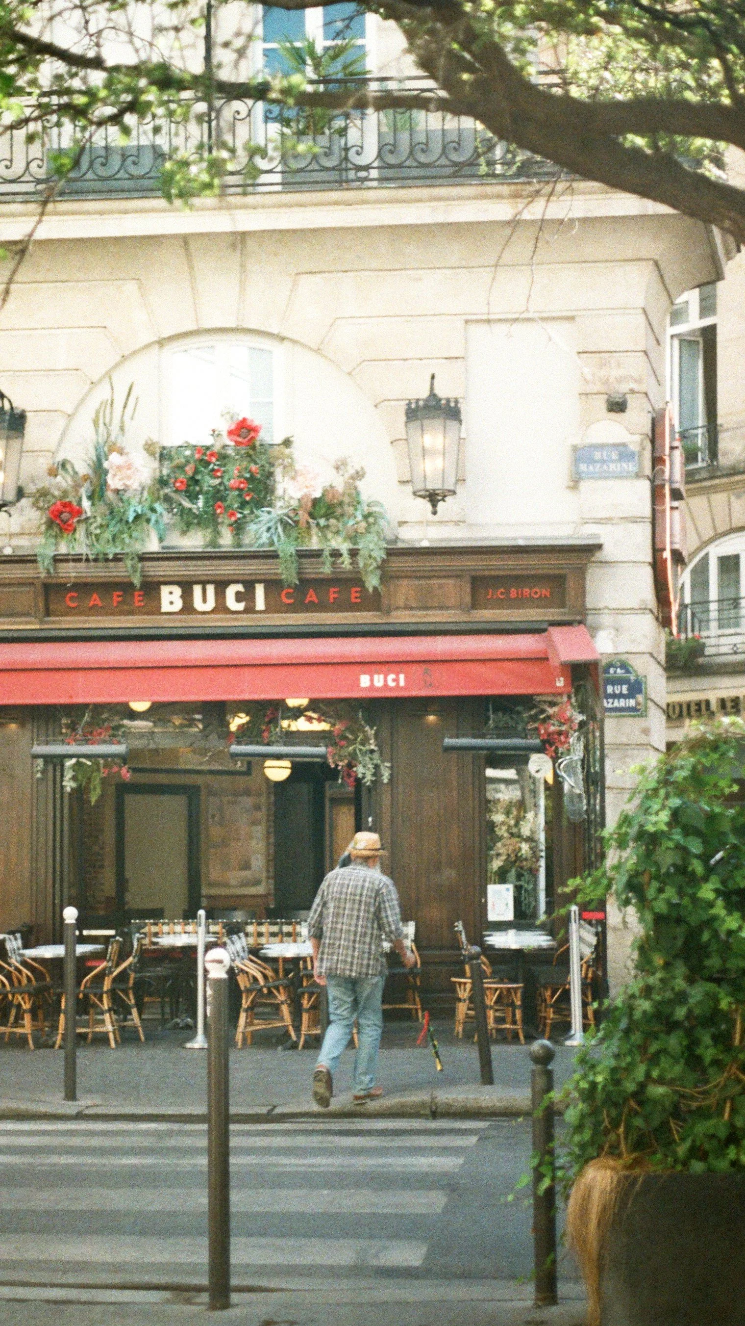 A street view of a Parisian cafe named 'Buci'. The cafe has outdoor seating with chairs and tables, and a red awning with the cafe's name. There are flower arrangements hanging around the entrance, and a man with a hat walking in front of the cafe.