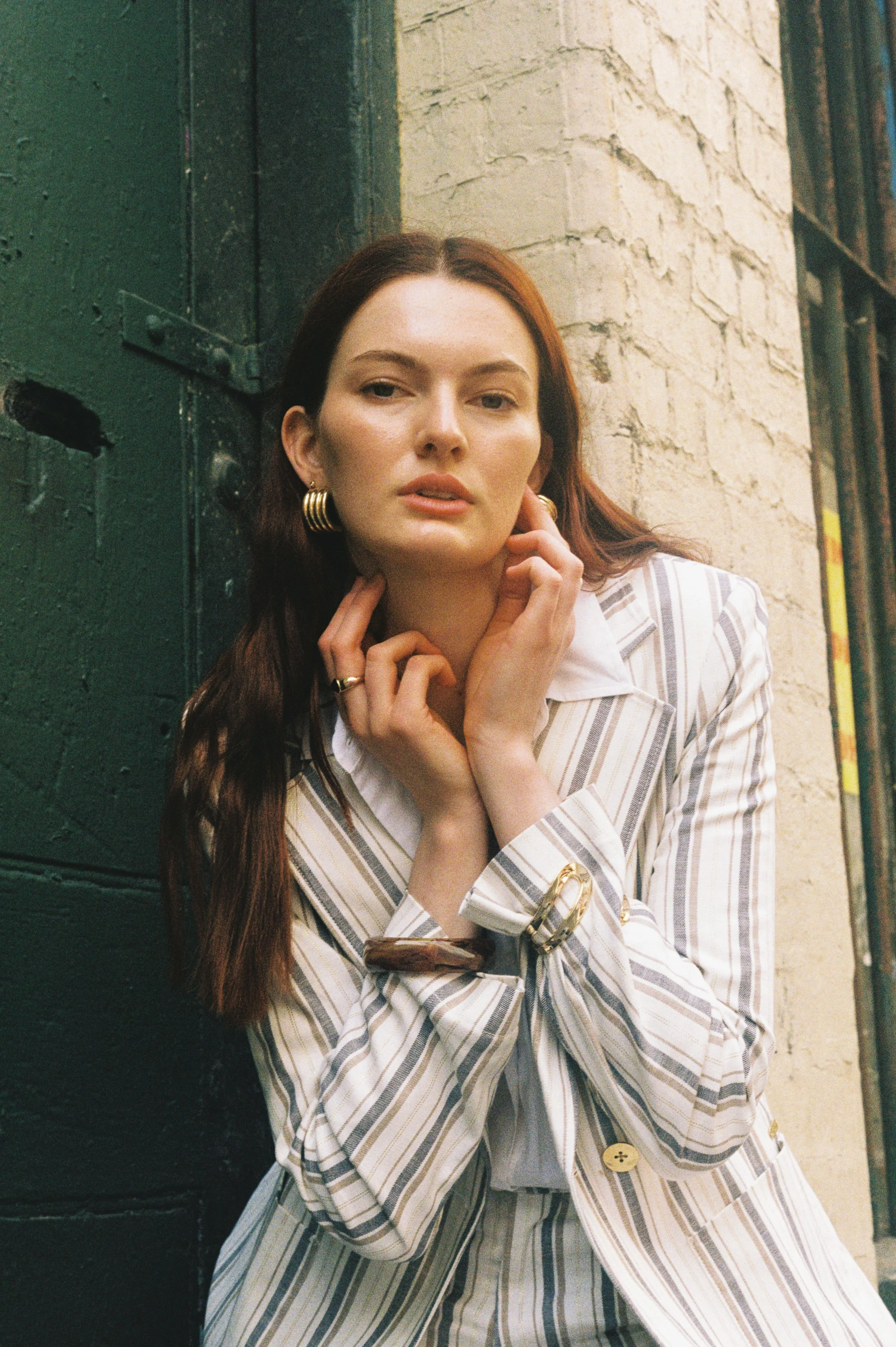 A woman with reddish-brown hair, wearing gold hoop earrings and a striped blazer, standing against a brick and green wall, looking at the camera with her hands gently touching her neck and chin.
