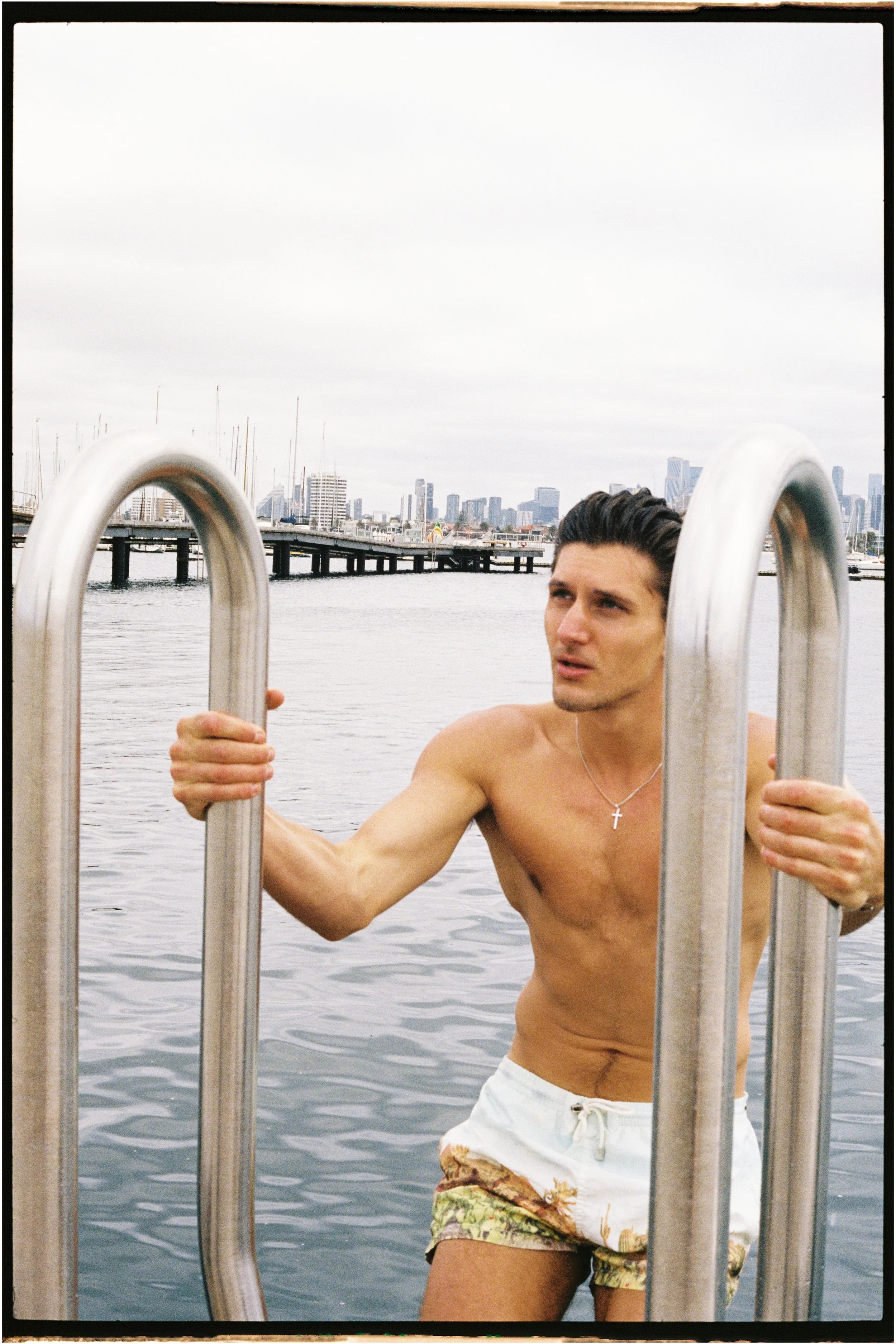 A shirtless man with dark hair and a cross necklace climbing up a metal ladder on a dock, with city skyline and water in the background.