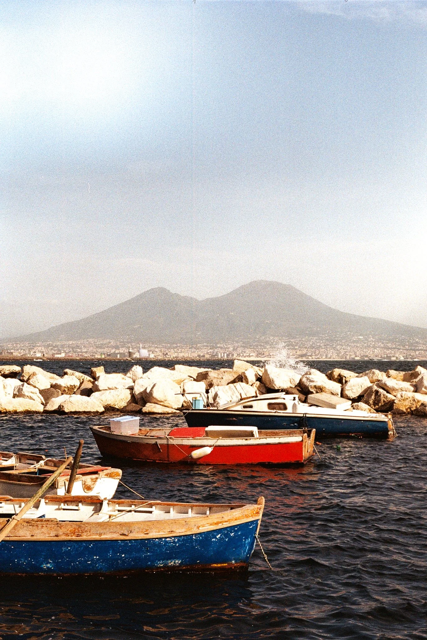 Boats anchored by a rocky shoreline with Mount Vesuvius in the background.