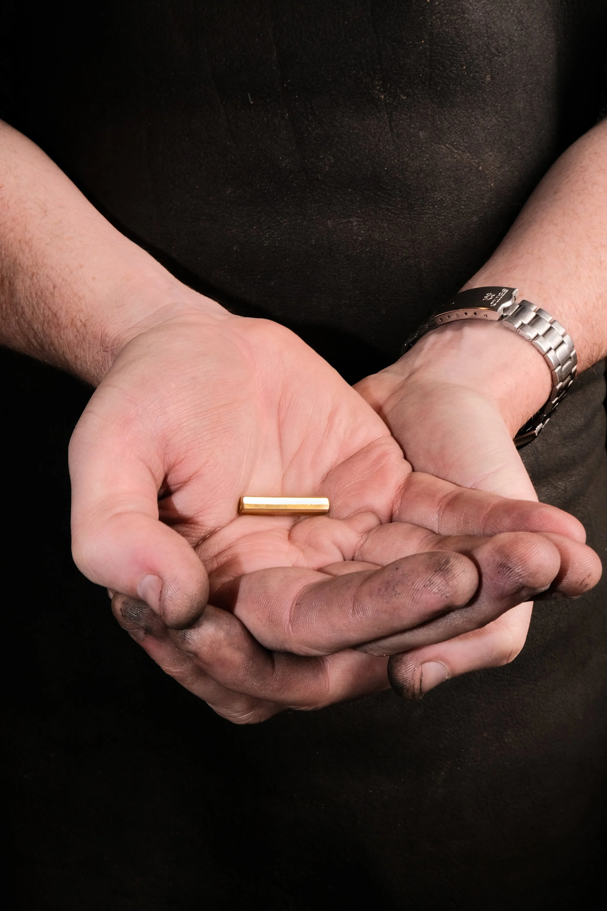 Two hands, one clean and one dirty, holding a small gold bar, with a person wearing a silver watch on their wrist.