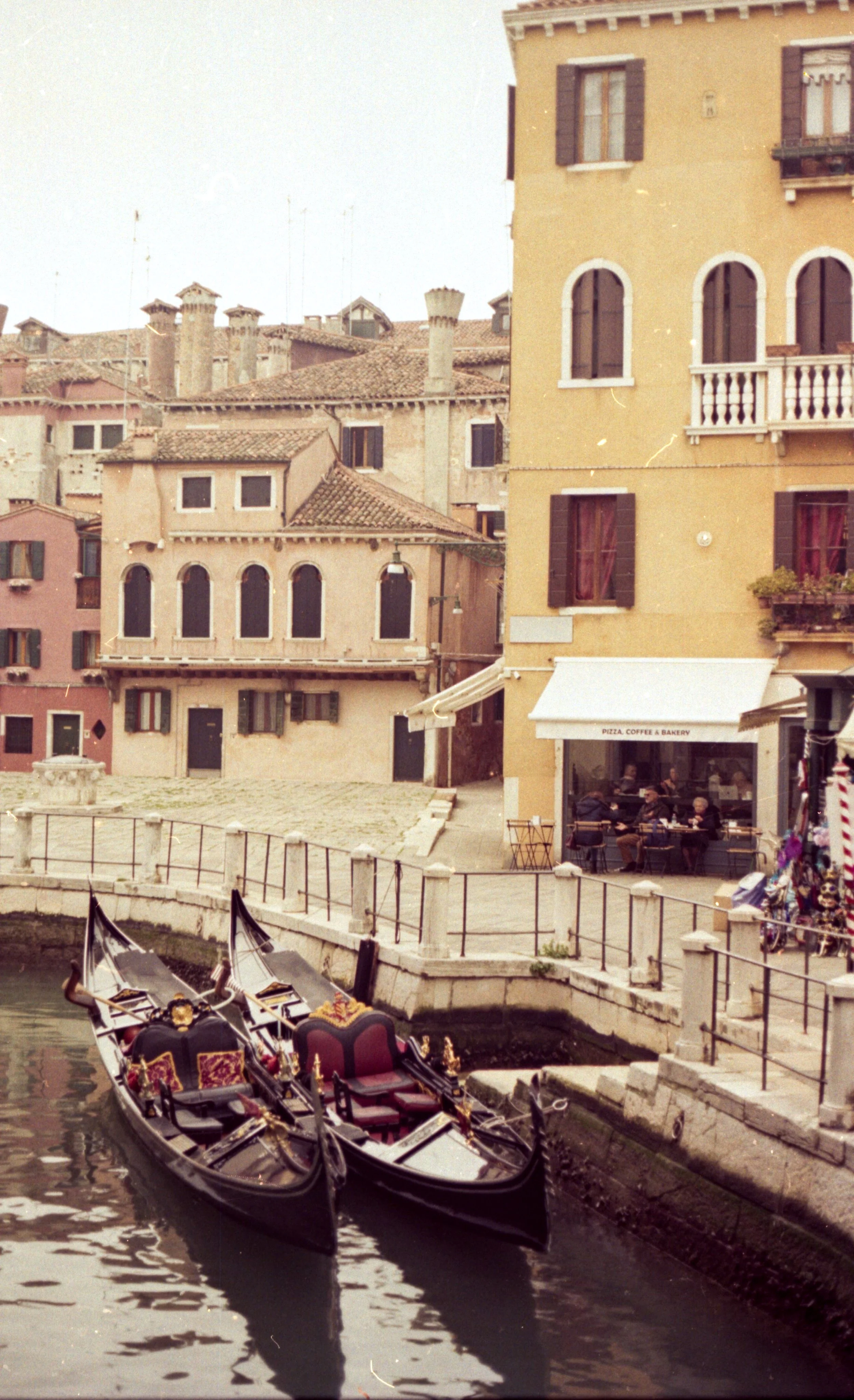 Venetian canal with two gondolas, colorful buildings, and a cafe with people dining outside.