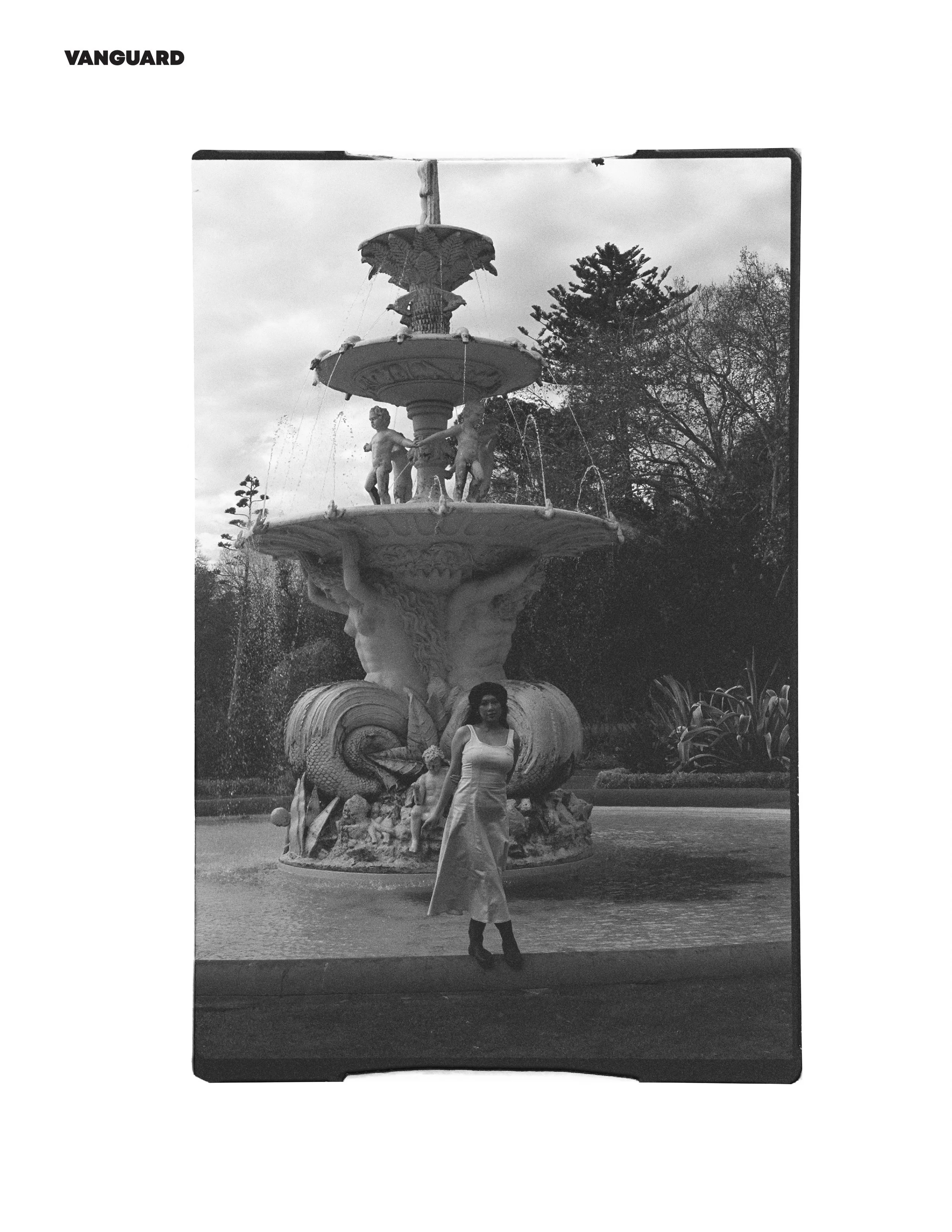 A black and white photo of a woman standing in front of a decorative fountain with statues of cherubs and lions, surrounded by trees and plants.