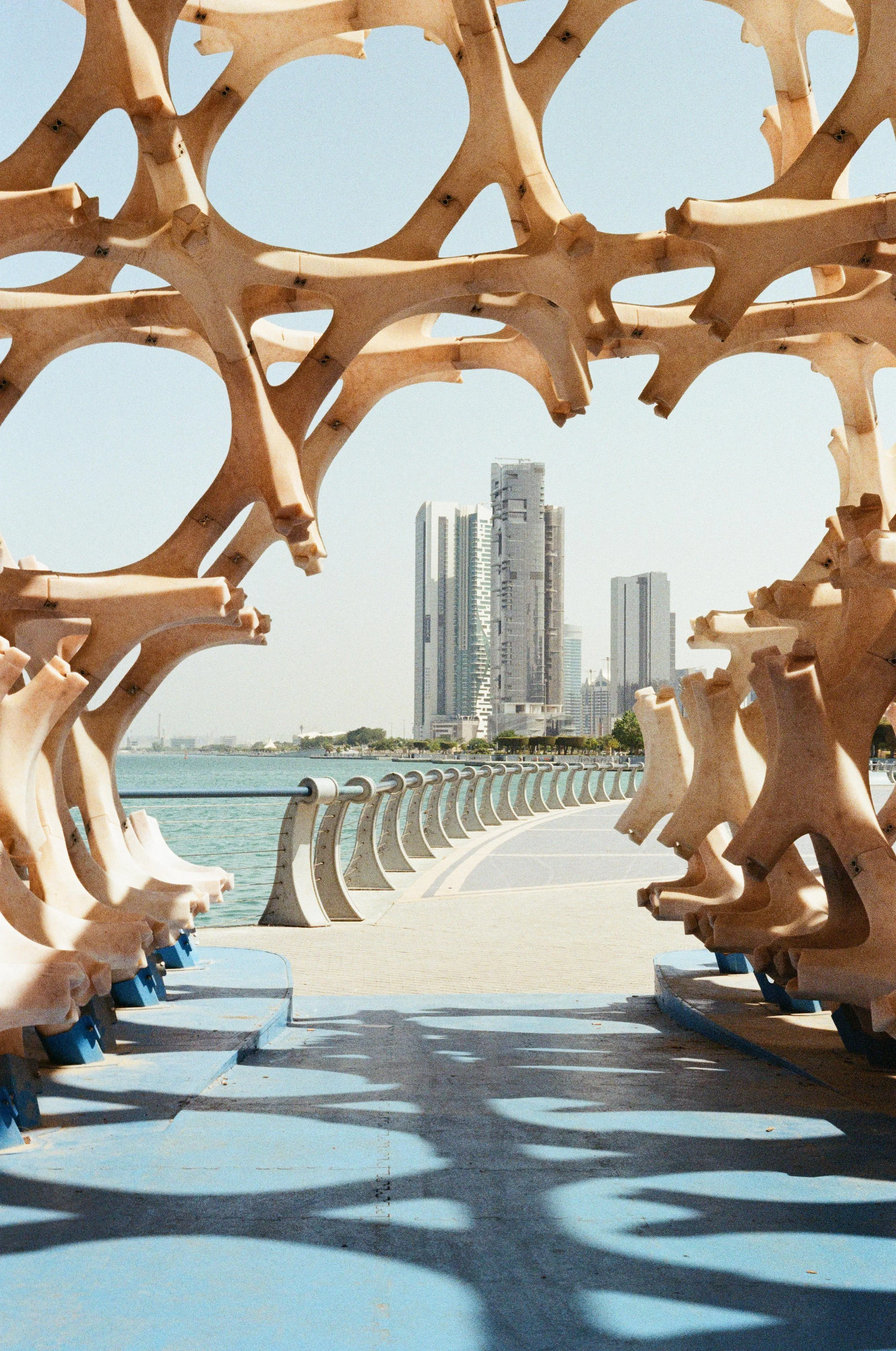 View through a wooden art installation of interconnected geometric shapes at a waterfront promenade, with skyscrapers and a city skyline in the background.