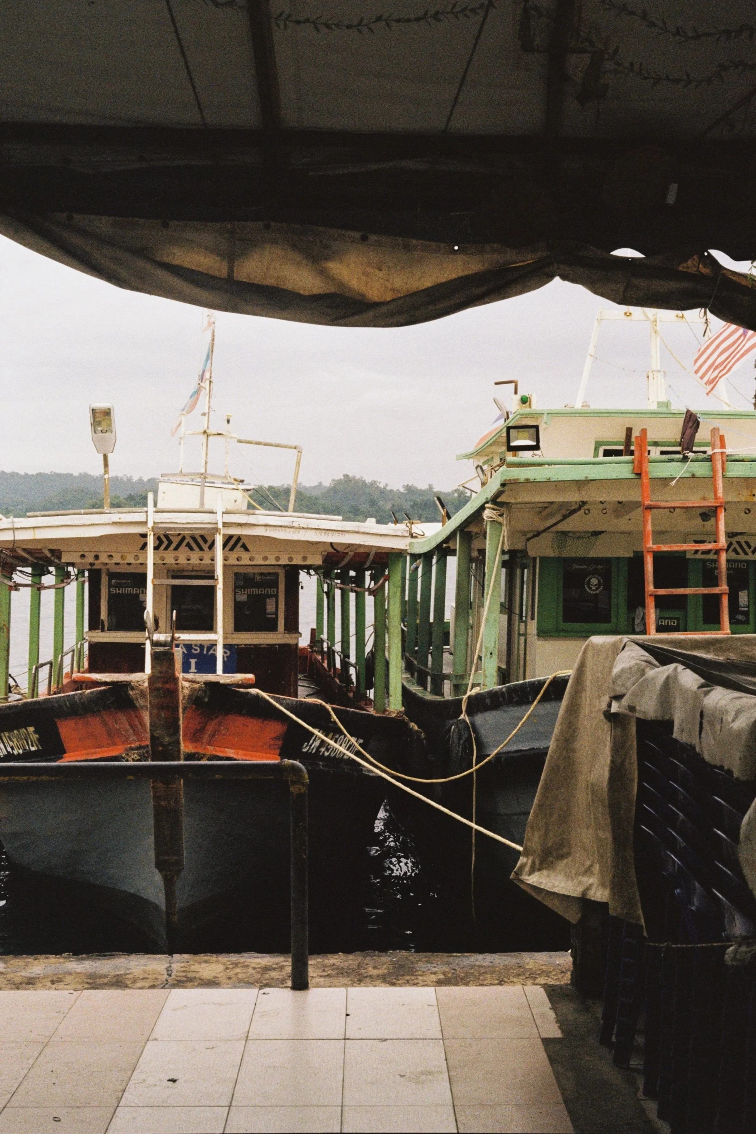 View from inside a dockside covered area showing boats docked at a marina, with hills and grey sky in the background.