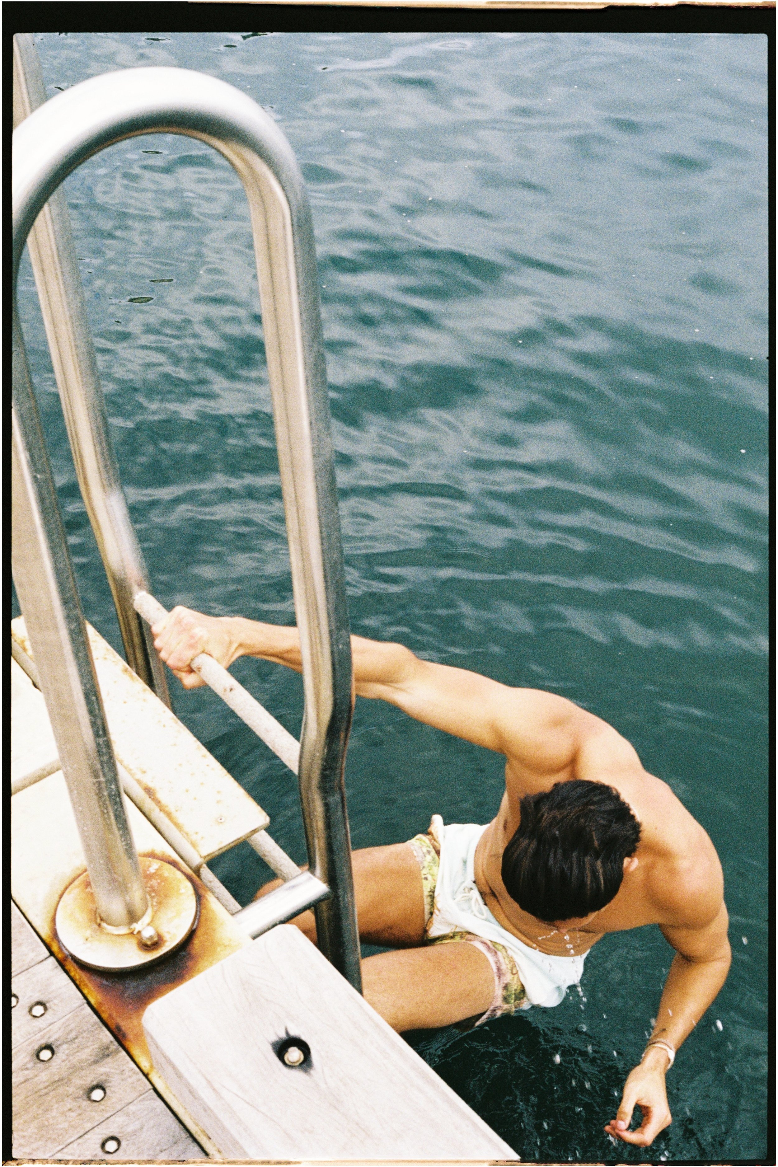 A man in floral swim shorts is swimming in a body of water, holding onto the ladder of a dock.