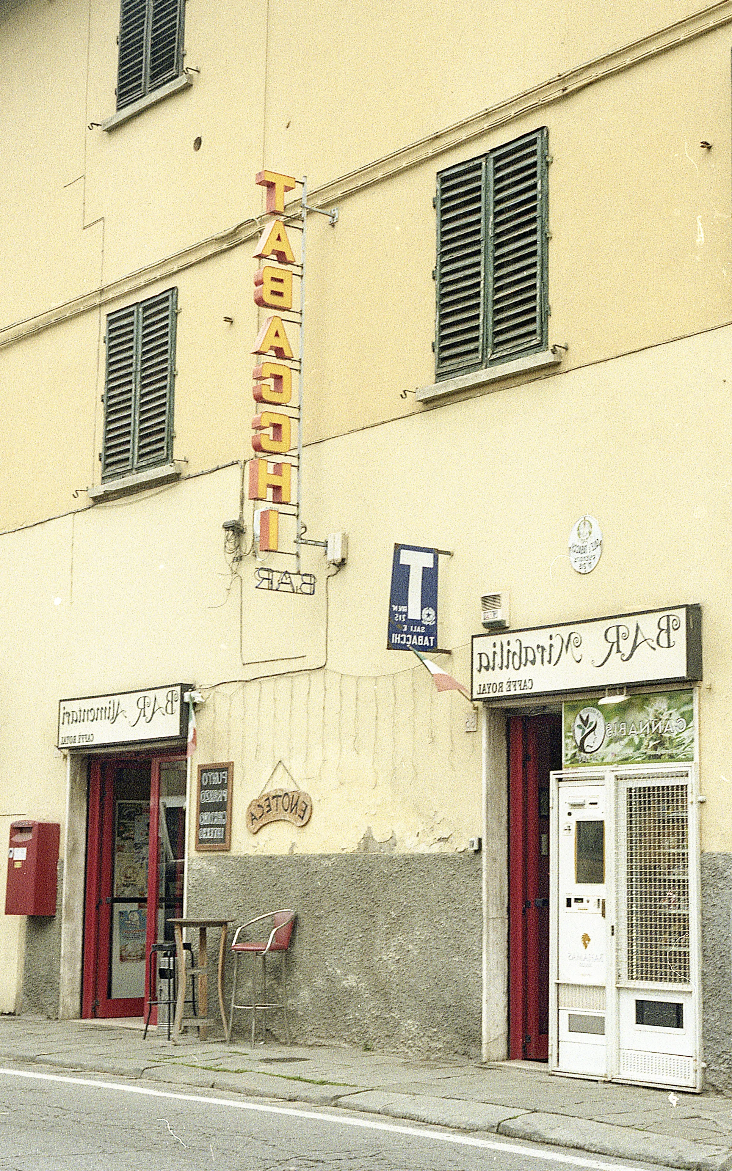 Exterior view of a building with a yellow facade, green window shutters, and signs for a restaurant or cafe and a bar, including a red mailbox, a small table with two chairs outside, and an automated vending or payment machine near the entrance.
