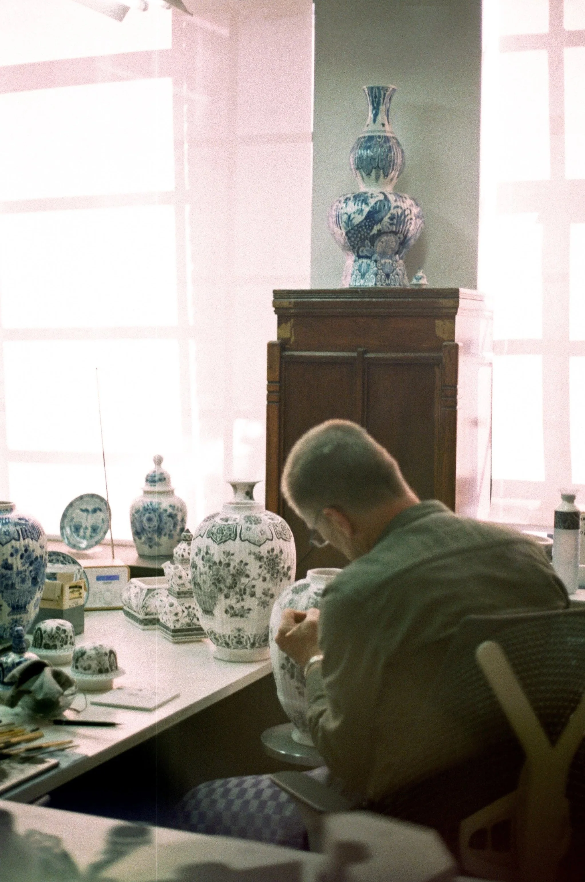 An artist working on intricate blue and white ceramic vases in a room with ample natural light coming through large windows.