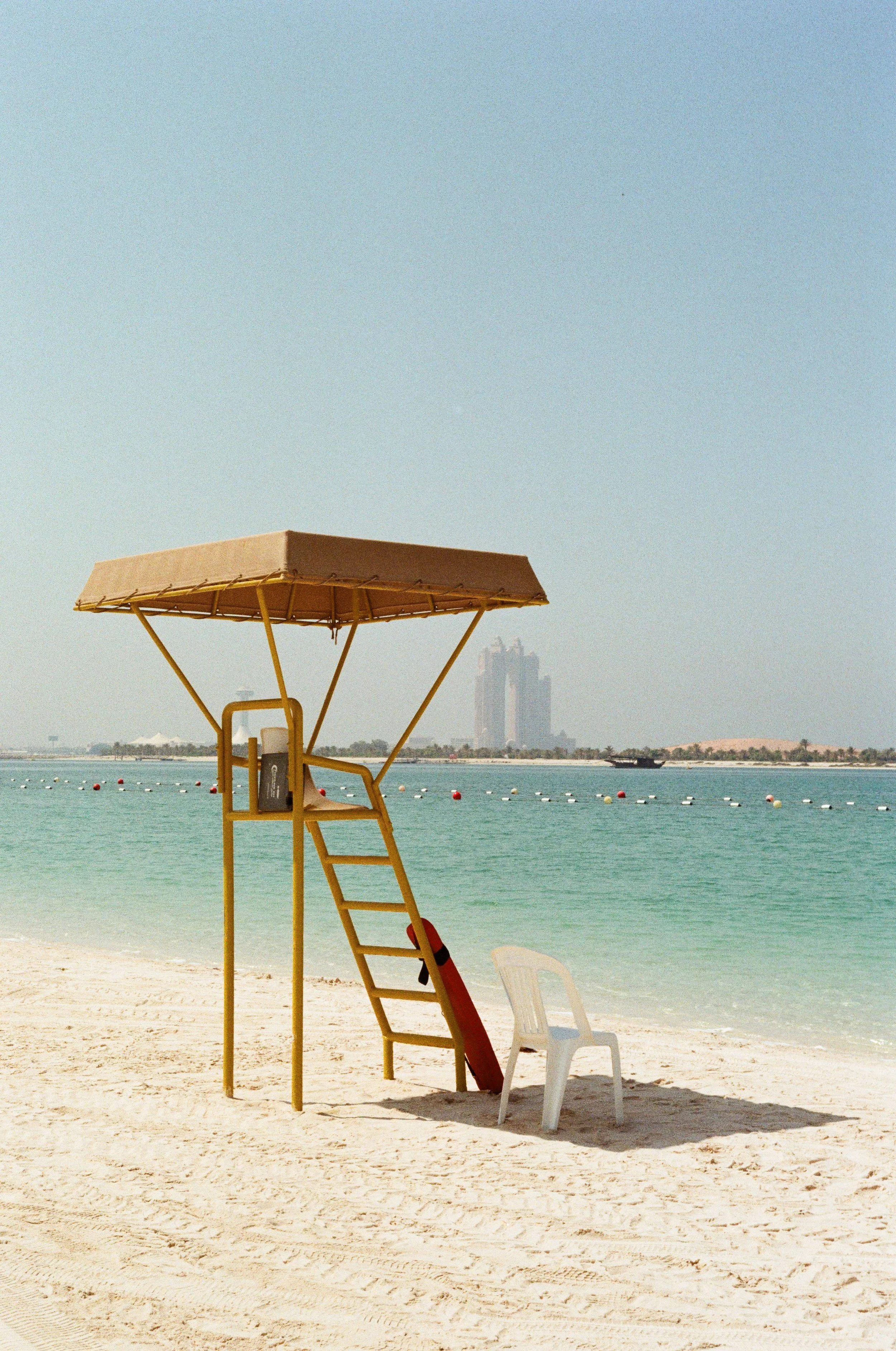 Empty lifeguard chair on a sandy beach with a city skyline in the background.