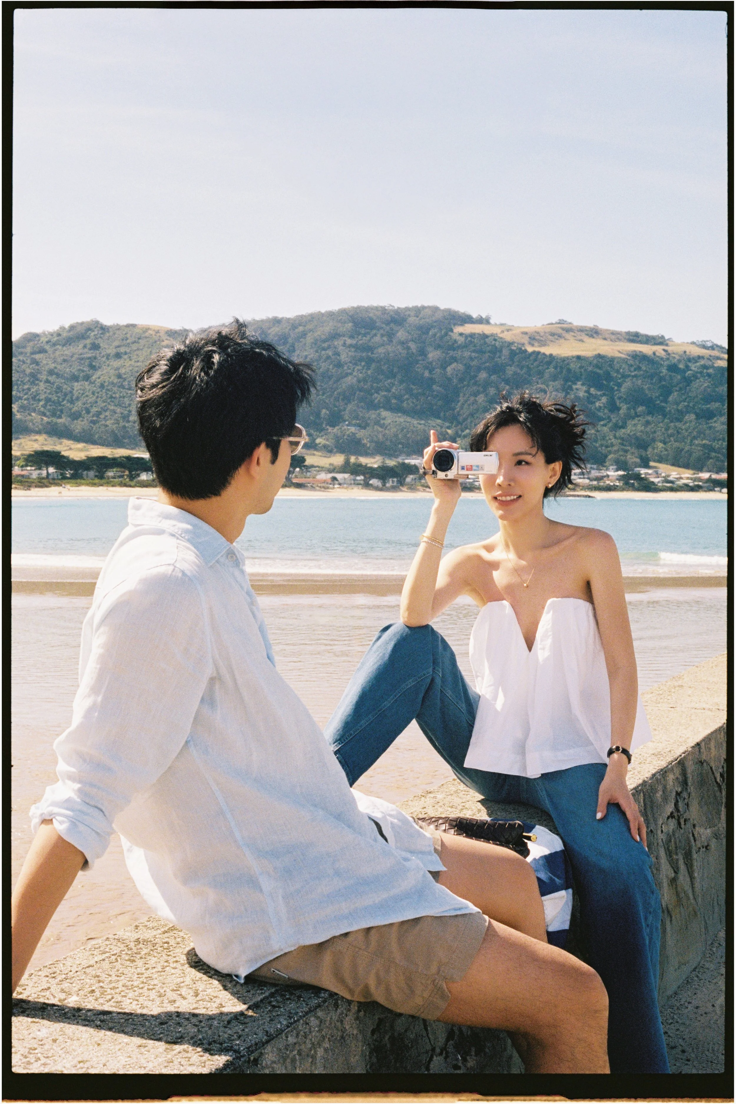 A young woman sitting on a stone ledge by the beach, holding a small camcorder and smiling as a young man sits nearby, all with a scenic mountain and water background.
