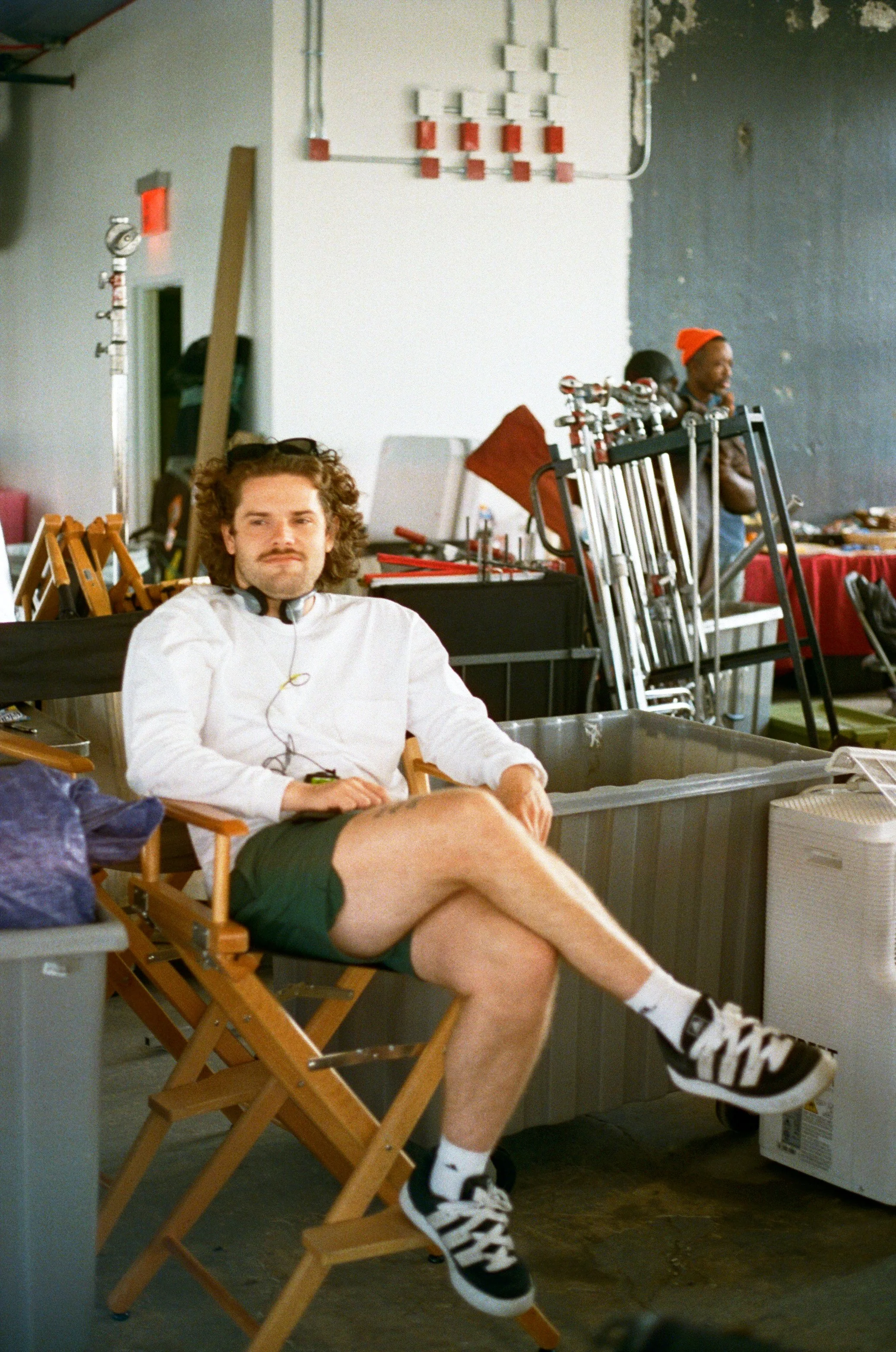 A man sitting on a director's chair with crossed legs, wearing a white long-sleeve shirt, black shorts, and sneakers, in what appears to be a film or photo shoot studio.