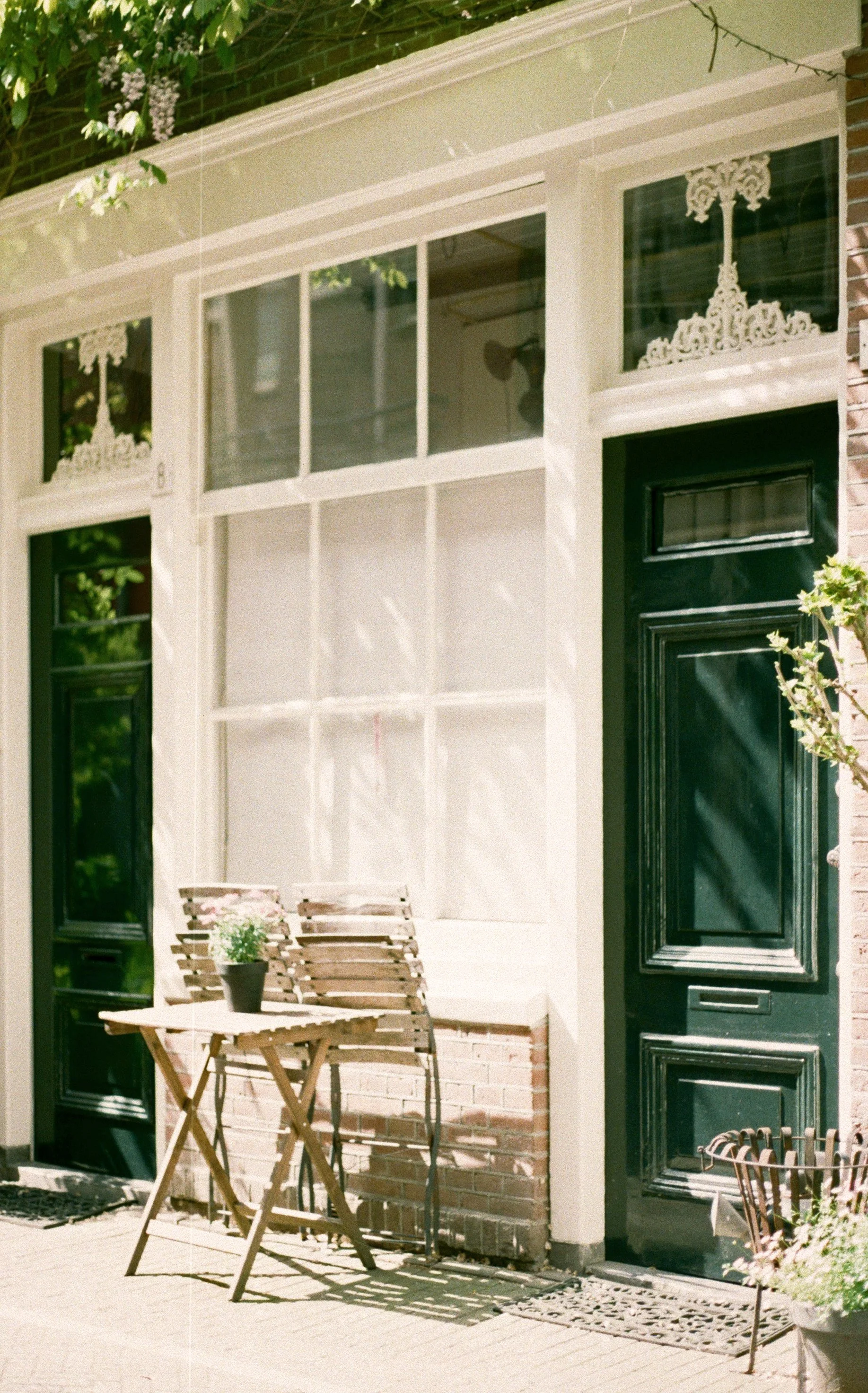 The exterior of a house with a green door, large window with frosted glass, and white decorative trim. A small wooden table with a potted plant and two wooden chairs are placed outside on a patio.