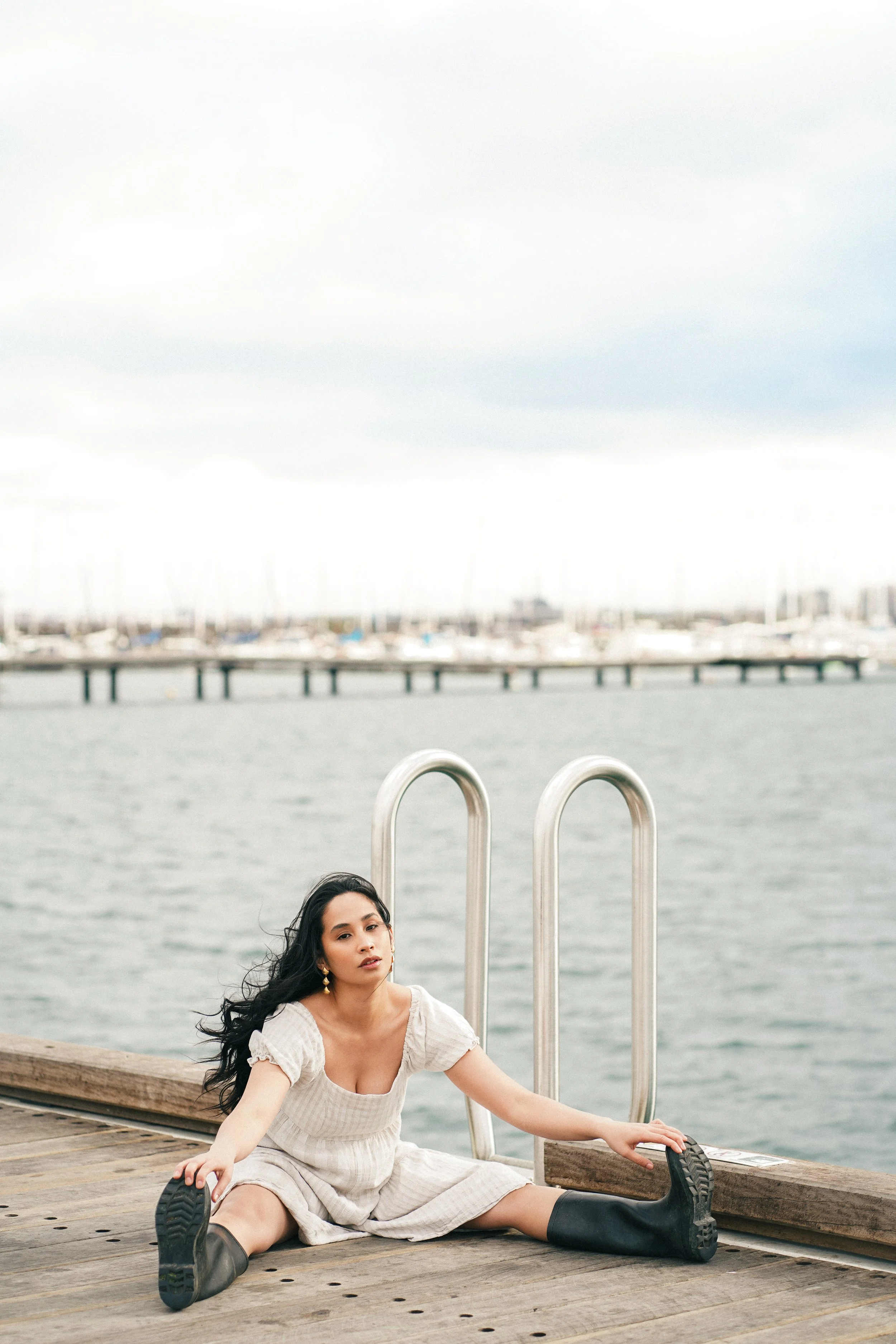 A young woman with long black hair posing on a wooden dock by the water, wearing a light-colored dress and black rain boots, with a marina and cloudy sky in the background.