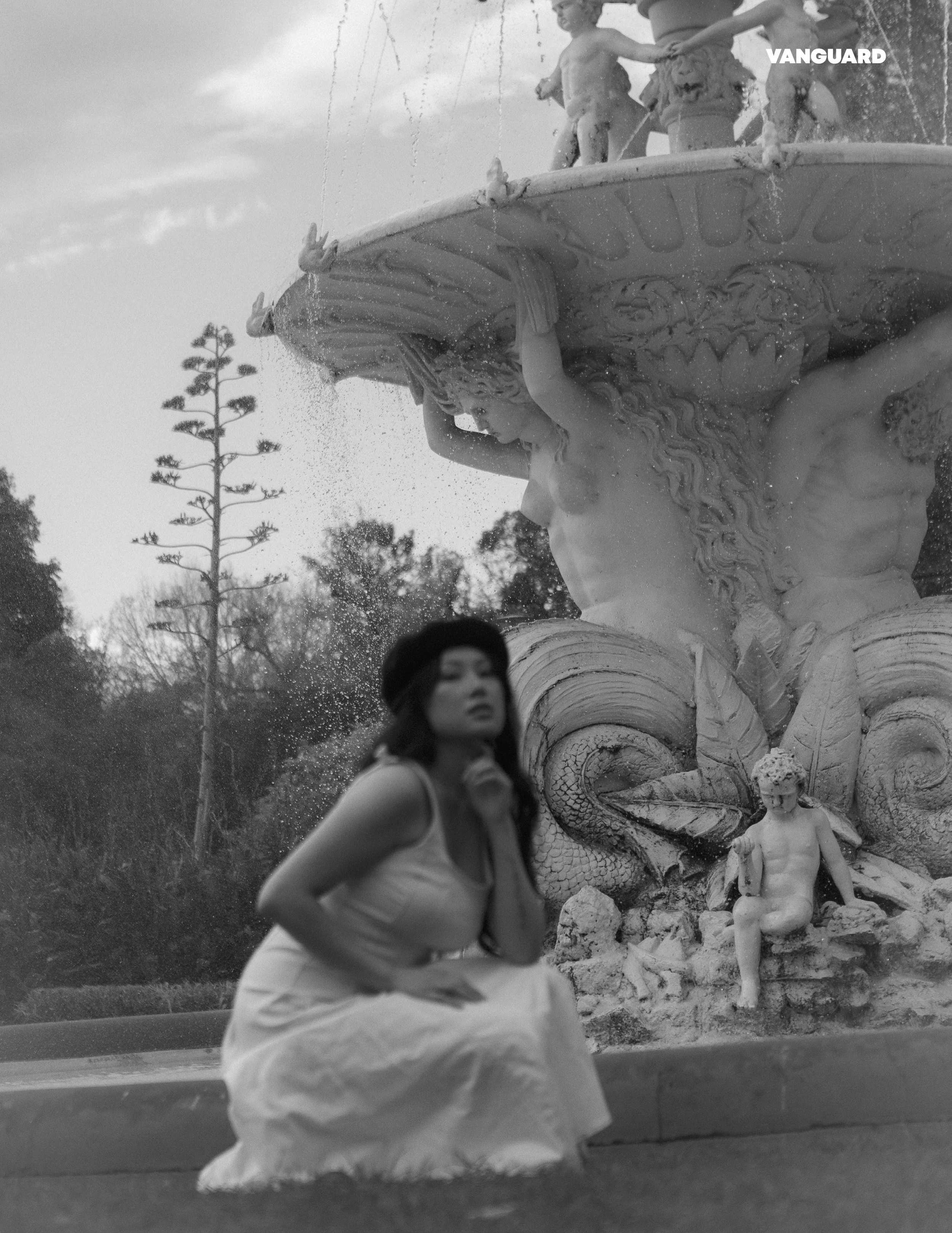 A woman sitting in front of a fountain with a sculpture of a mermaid and children, with trees in the background. The photo is in black and white.
