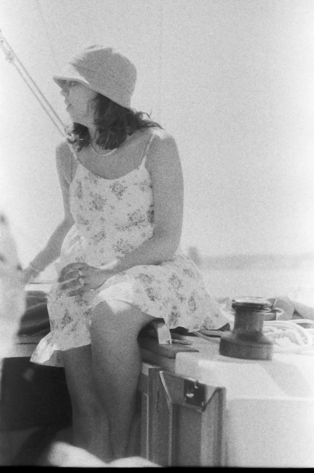 Black and white photo of a woman sitting on a wooden surface, wearing a floral dress and a hat, with a spool of thread nearby.
