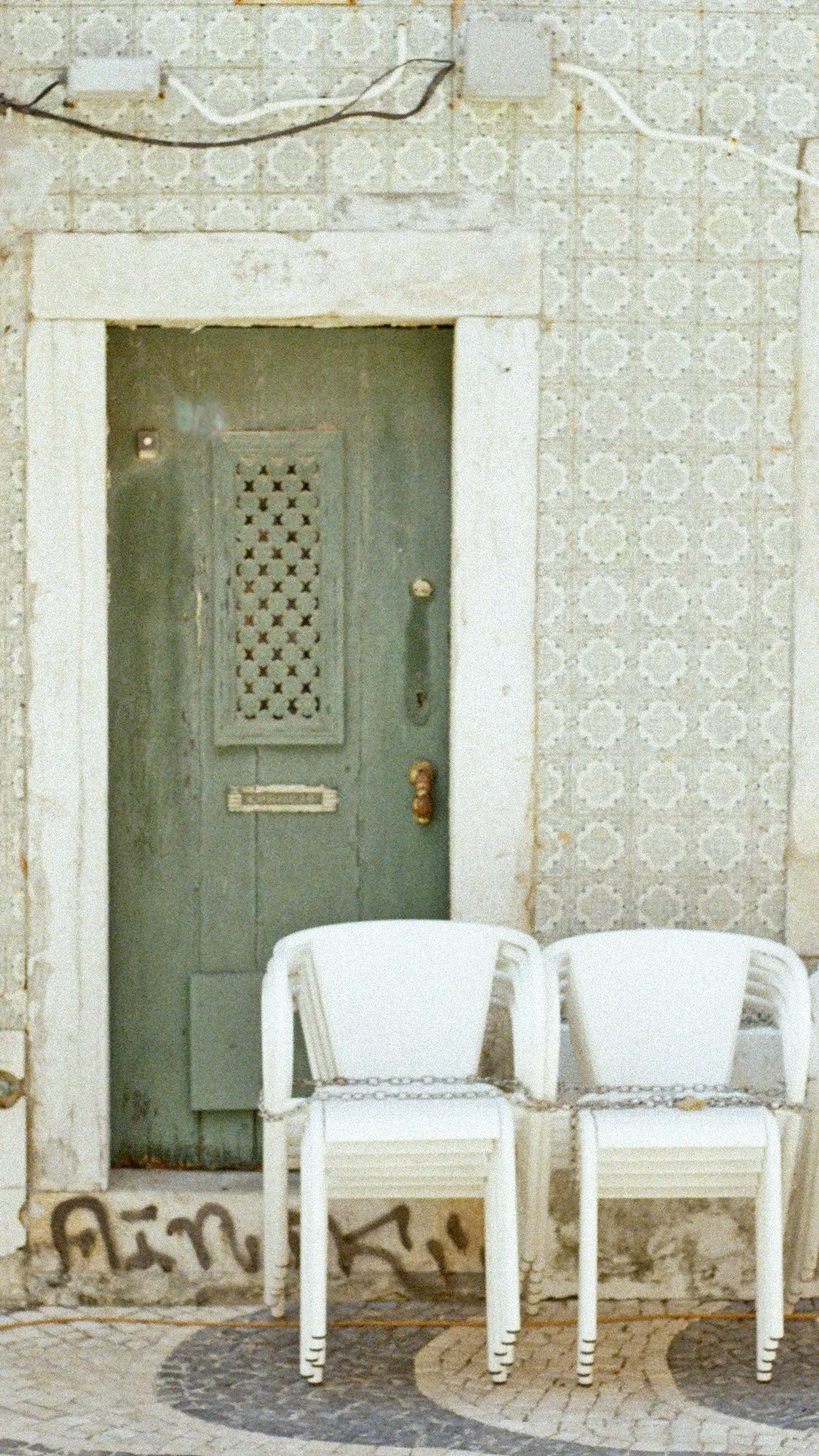 Two white chairs chained together in front of an old green door with peeling paint, set in an aged wall with patterned tile.